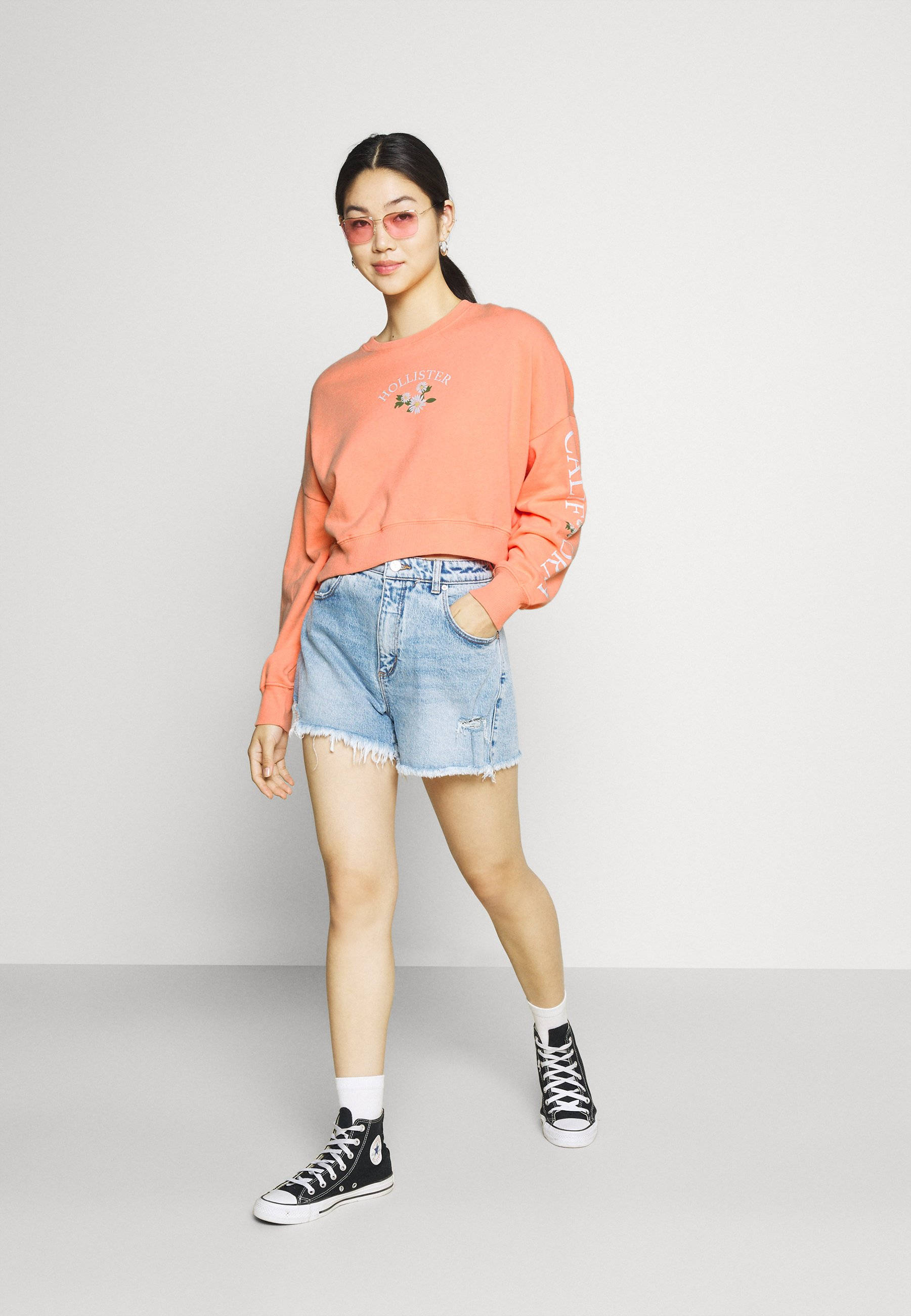 Hollister 桌布 壁紙 — 一位身穿珊瑚色運動衫和牛仔短褲的年輕女性，在純白色工作室背景前自信地擺姿勢。