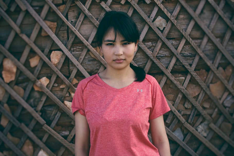 粉紅色 桌布 壁紙 — 一位身穿珊瑚色運動衫的年輕女子冷靜地站在帶有石頭裝飾的質樸木格柵牆前。