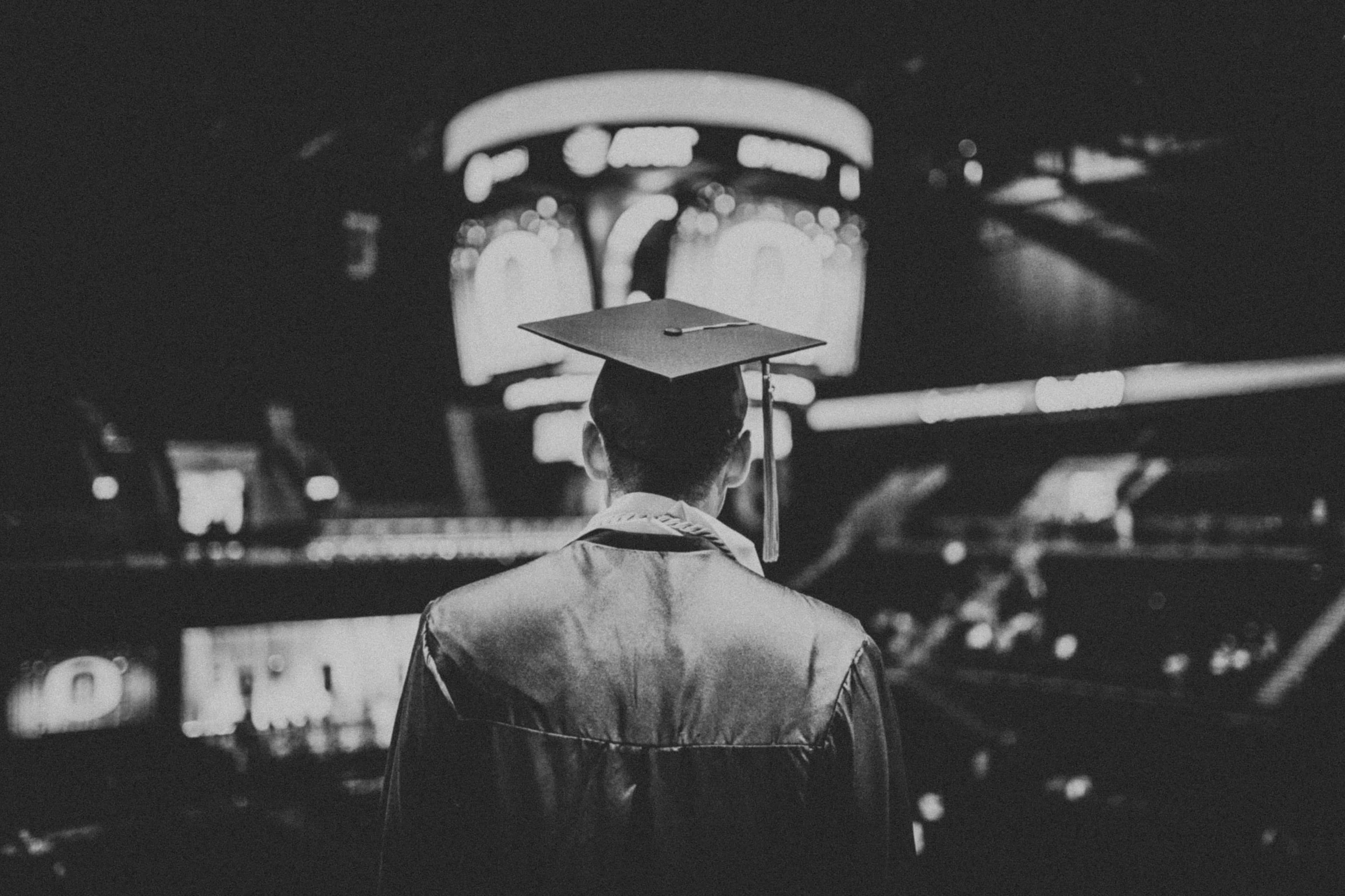 Graduation 桌布 壁紙 — 一位身穿畢業袍和學士帽的畢業生夜晚獨自站在體育館，凝視著明亮的記分板。