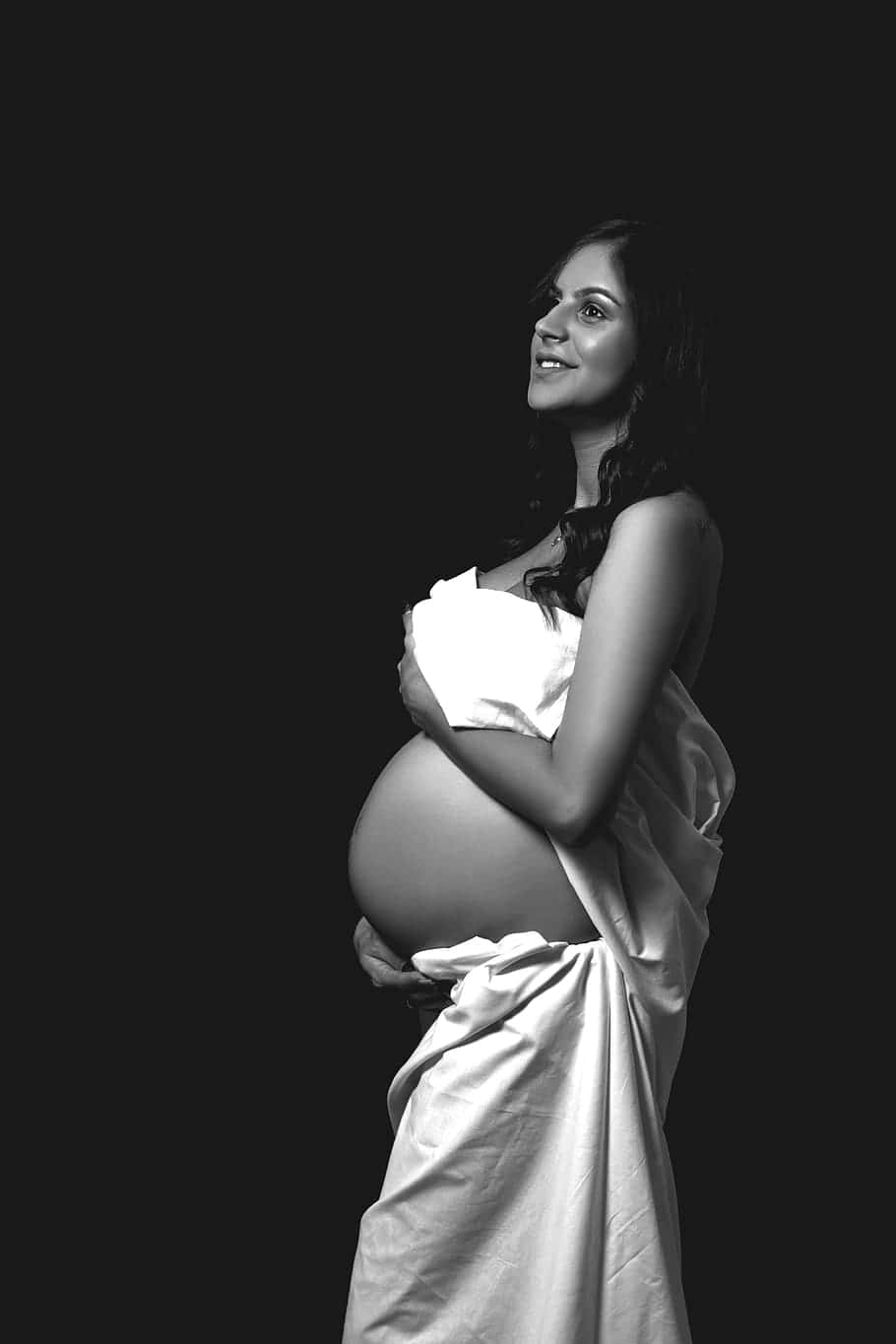 pregnancy 桌布 壁紙 — 一位身穿白布的孕婦在純黑背景前輕輕微笑，懷抱著自己的肚子。