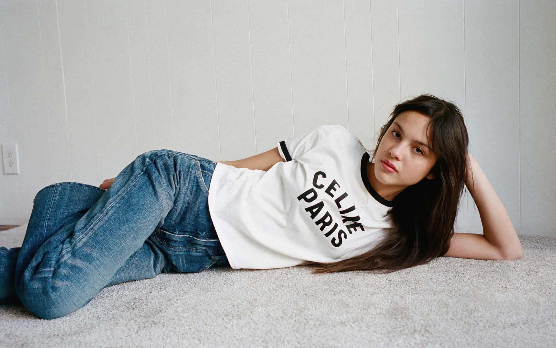 奧莉維亞·羅德里戈 桌布 壁紙 — 一位身穿白色 'CELINE PARIS' T恤和藍色牛仔褲的年輕女性，在淺色木鑲板牆邊的地毯上休息。