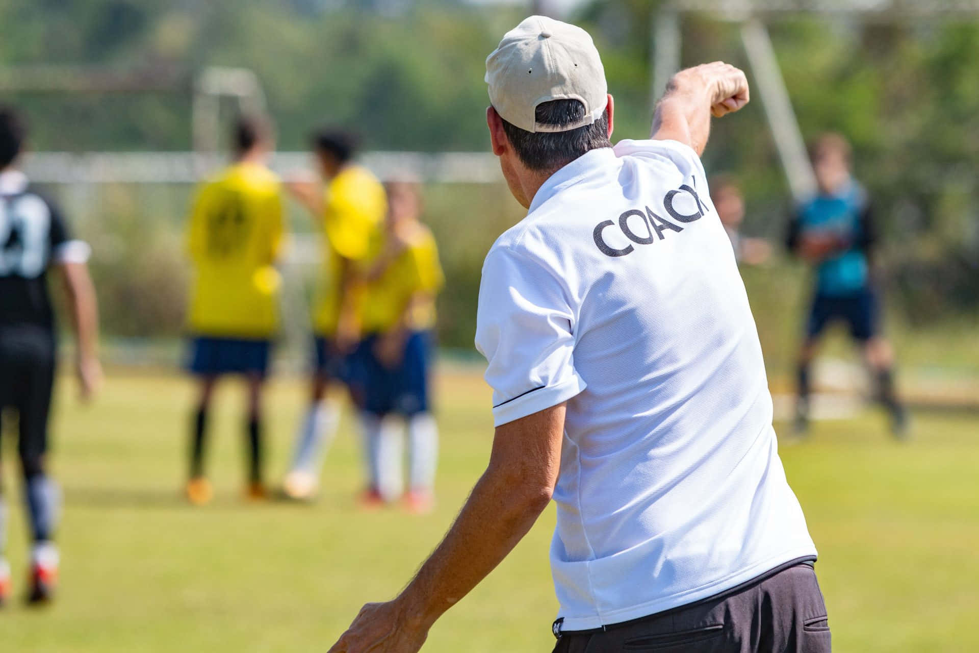 Coach 桌布 壁紙 — 一位身穿白色 'COACH' 上衣的足球教練在陽光明媚的草地上用力做手勢，身後有身穿黃色球衣的球員在訓練。
