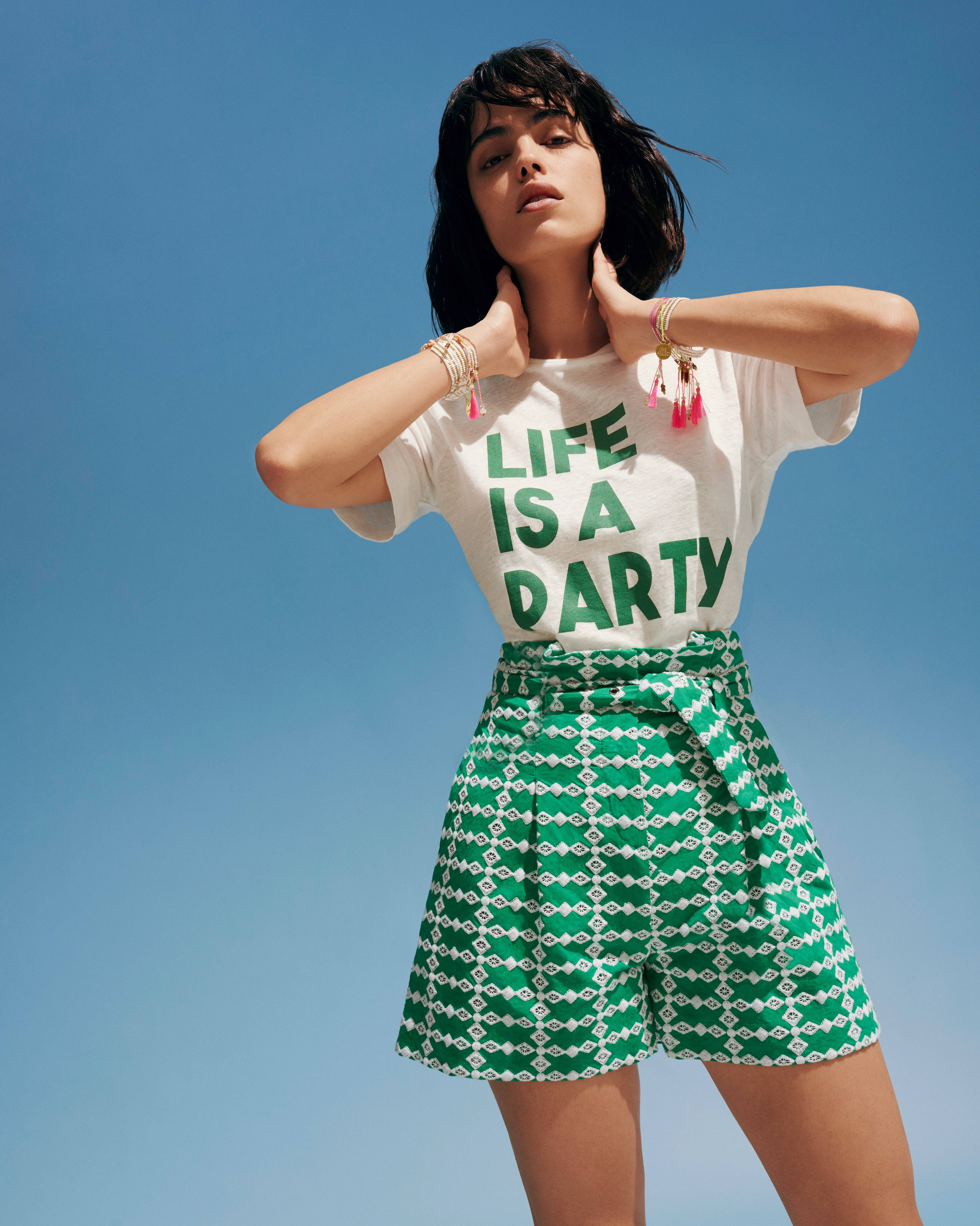 時尚 桌布 壁紙 — 一位身穿白色「LIFE IS A PARTY」T恤和綠色圖案短褲的女性，在清澈的藍天下自信地擺姿勢。