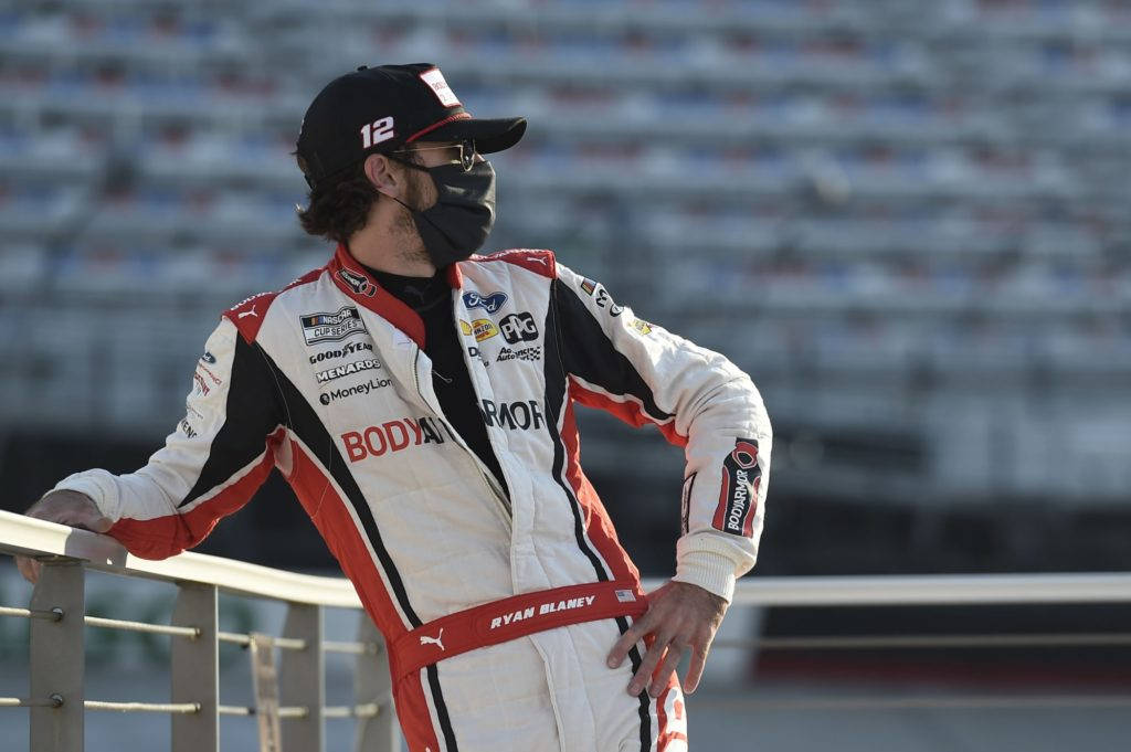 Ryan Blaney 桌布 壁紙 — 一位身穿白色、紅色和黑色賽車服的NASCAR車手在明亮的日光下靠在空蕩蕩的體育場的欄杆上。