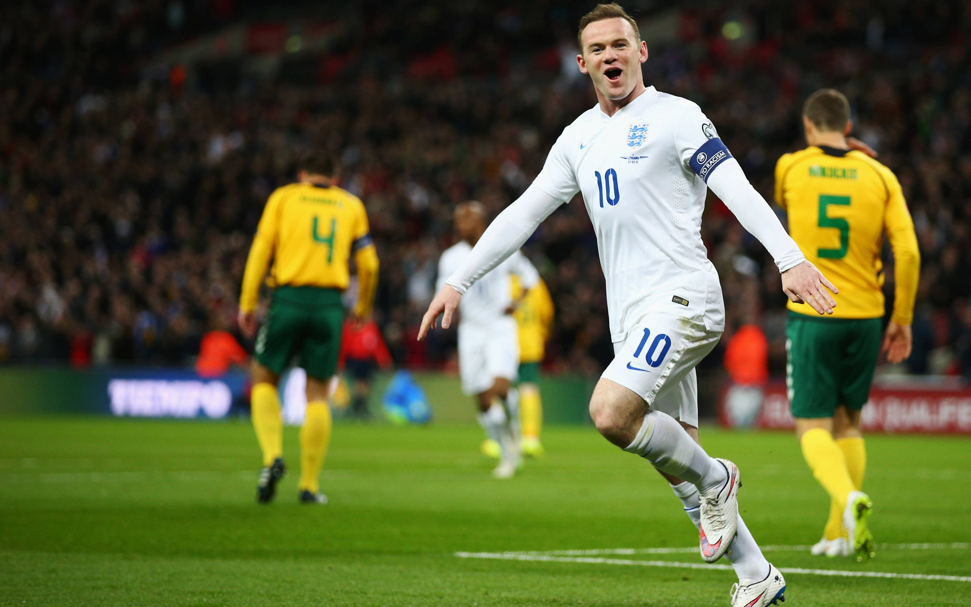 Rooney 桌布 壁紙 — 一位身穿白色10號球衣的英格蘭足球員在充滿活力的綠色球場上慶祝進球，背景是身穿黃綠色球衣的模糊對手。