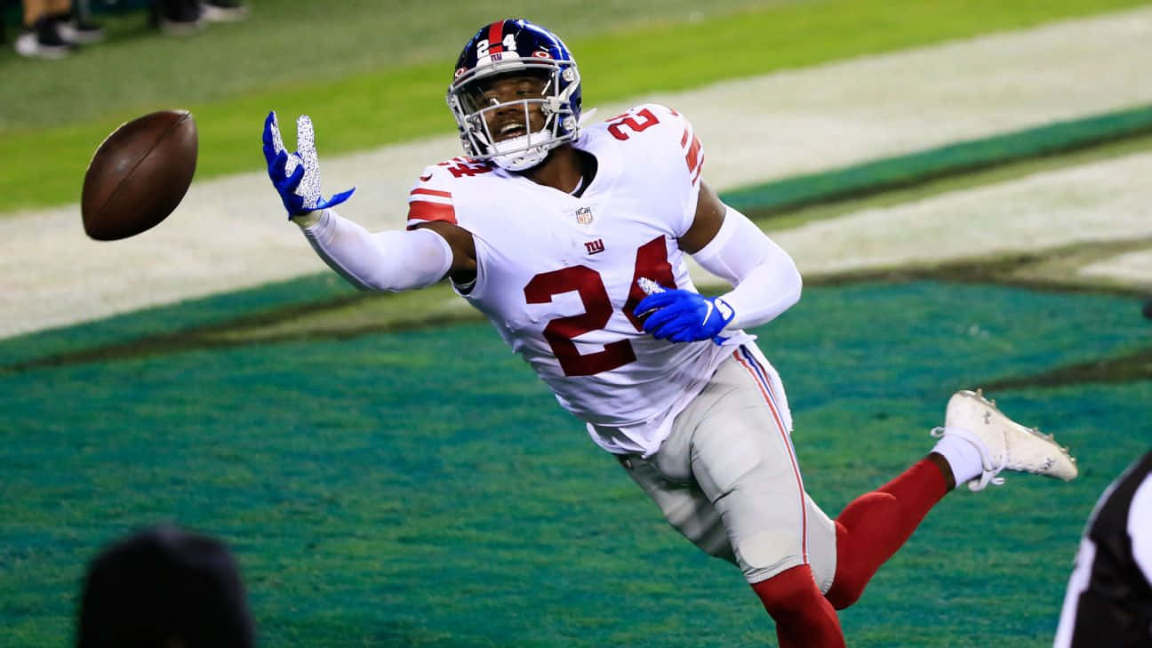James Bradberry 桌布 壁紙 — 一位身穿白色24號球衣的紐約巨人隊美式足球員，戴著藍色手套在空中跳躍，伸手去接綠色球場上的一個棕色美式足球。