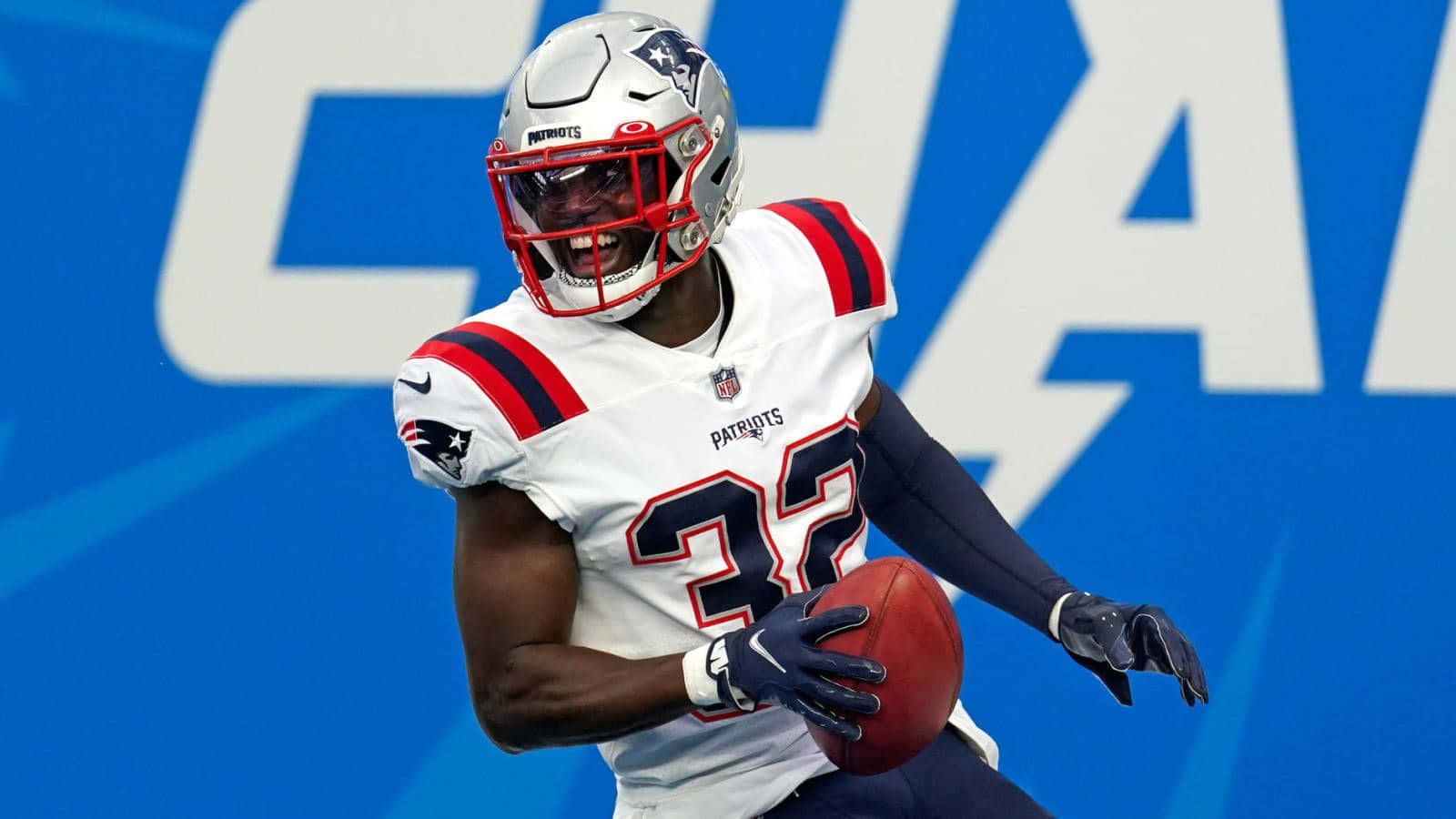 Devin Mccourty 桌布 壁紙 — 一位身穿白色32號球衣的新英格蘭愛國者隊美式足球員，在帶有大型白色字母的藍色背景上持球奔跑。