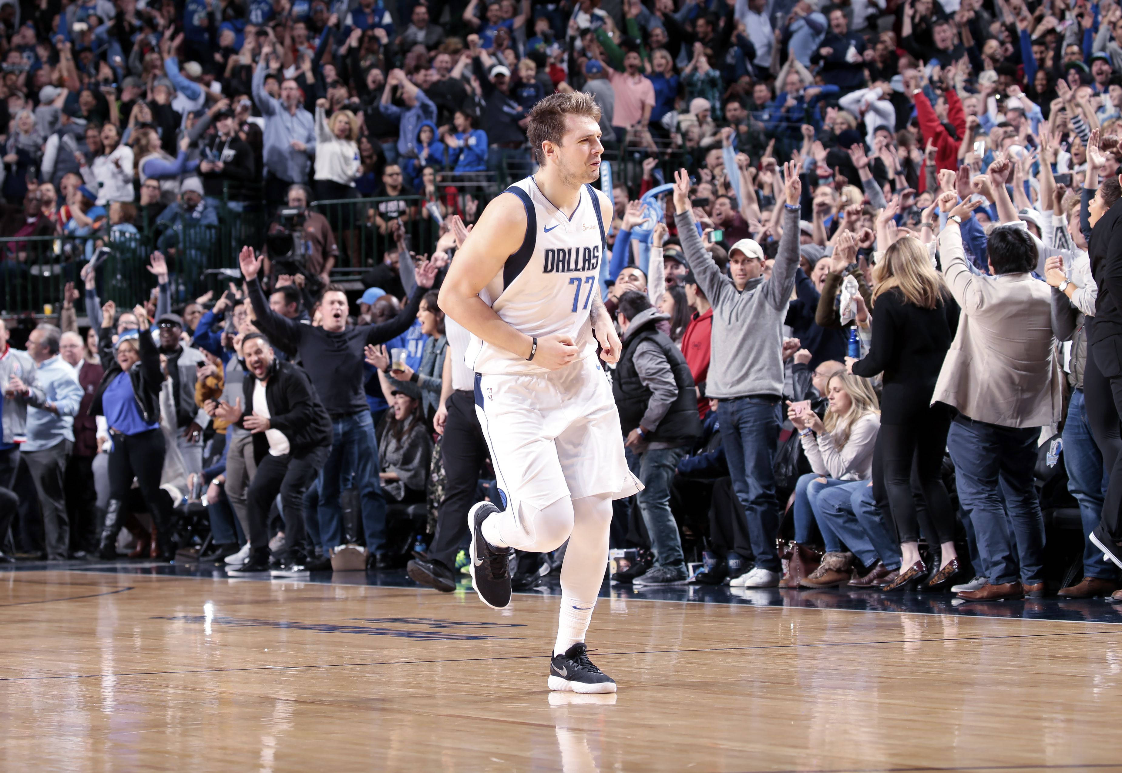 4K NBA 桌布 壁紙 — 一位身穿白色77號球衣的達拉斯獨行俠籃球運動員在球場上歡快地奔跑，而咆哮的球迷們歡呼並舉起雙手。