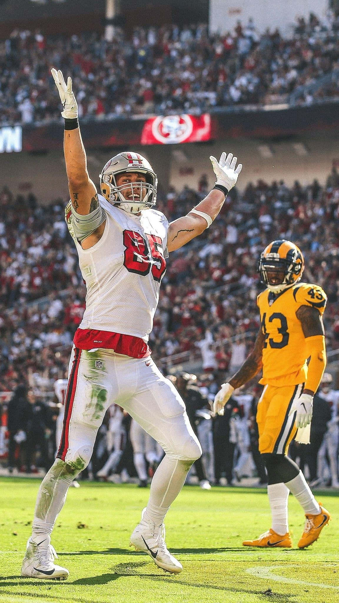 Football Players 桌布 壁紙 — 一位身穿白色85號球衣的舊金山49人美式足球員在陽光明媚的球場上高舉雙臂慶祝勝利，而一位身穿黃色球衣的匹茲堡鋼人球員則在附近走過。