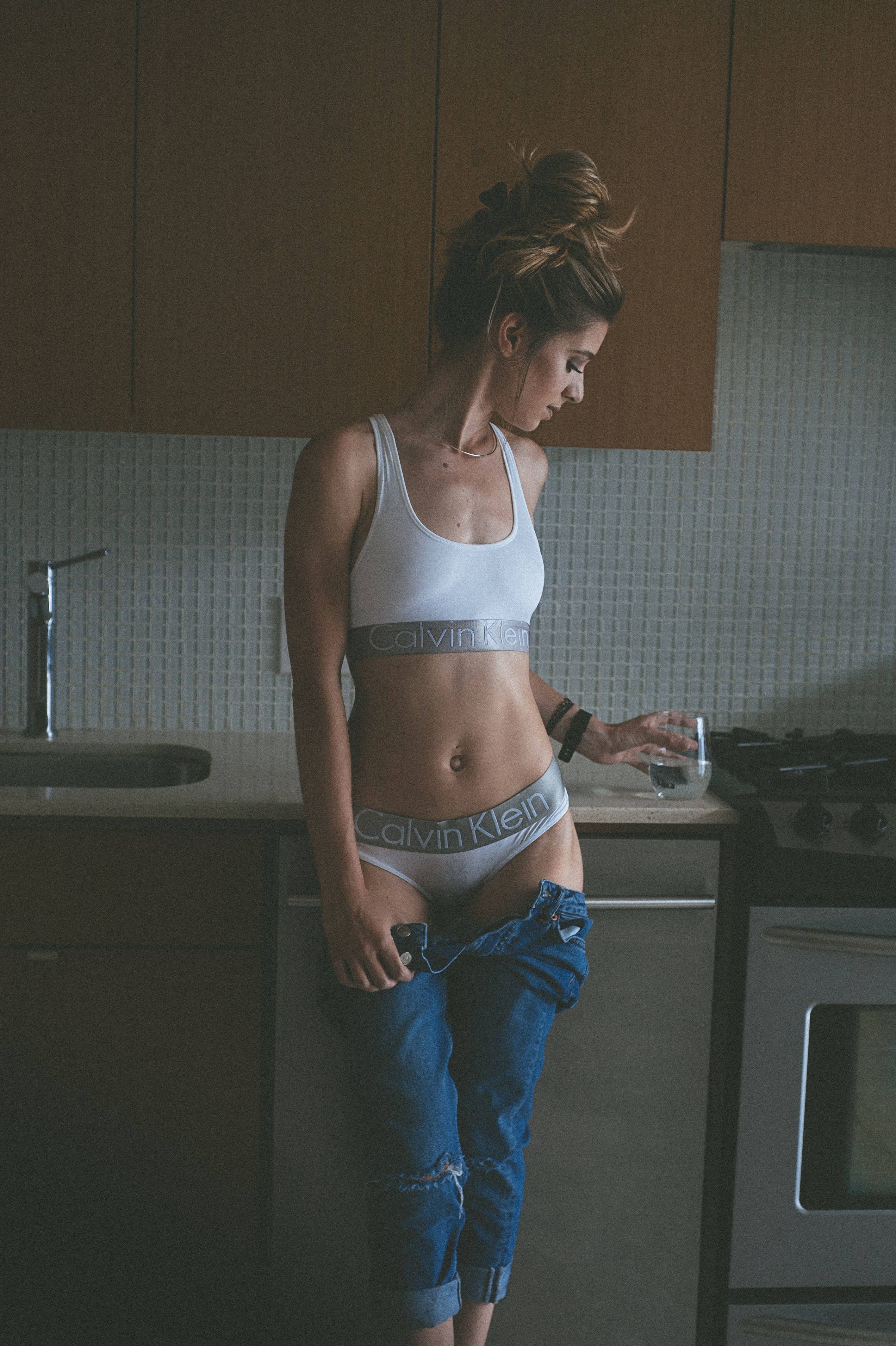 hot woman 桌布 壁紙 — 一位身穿白色Calvin Klein內衣和破洞牛仔褲的女性，站在現代化廚房裡，手持一杯水。