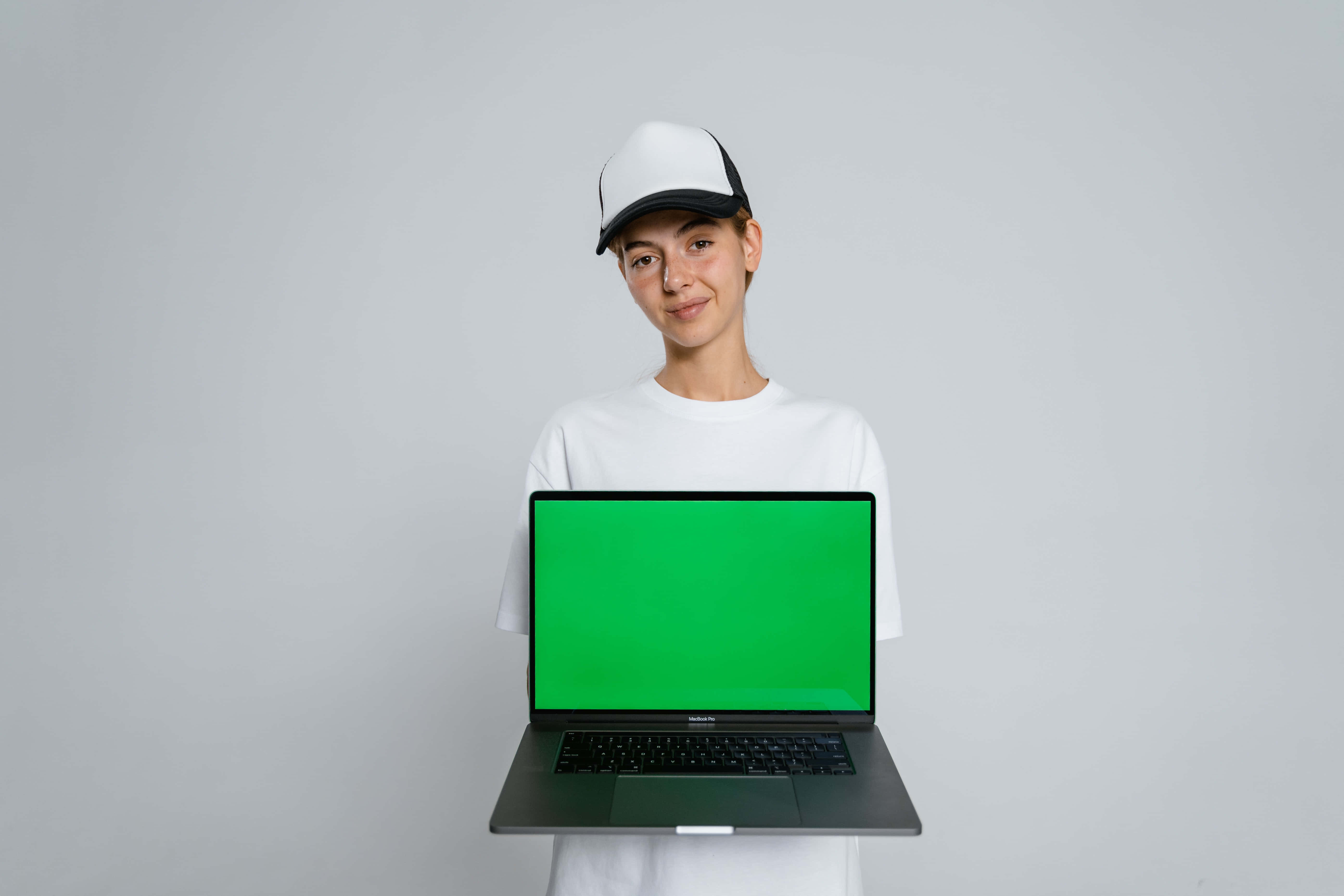 筆電 桌布 壁紙 — 一位身穿白色T恤和帽子的年輕女性，在純灰色背景前拿著一台帶有綠幕的筆記型電腦。