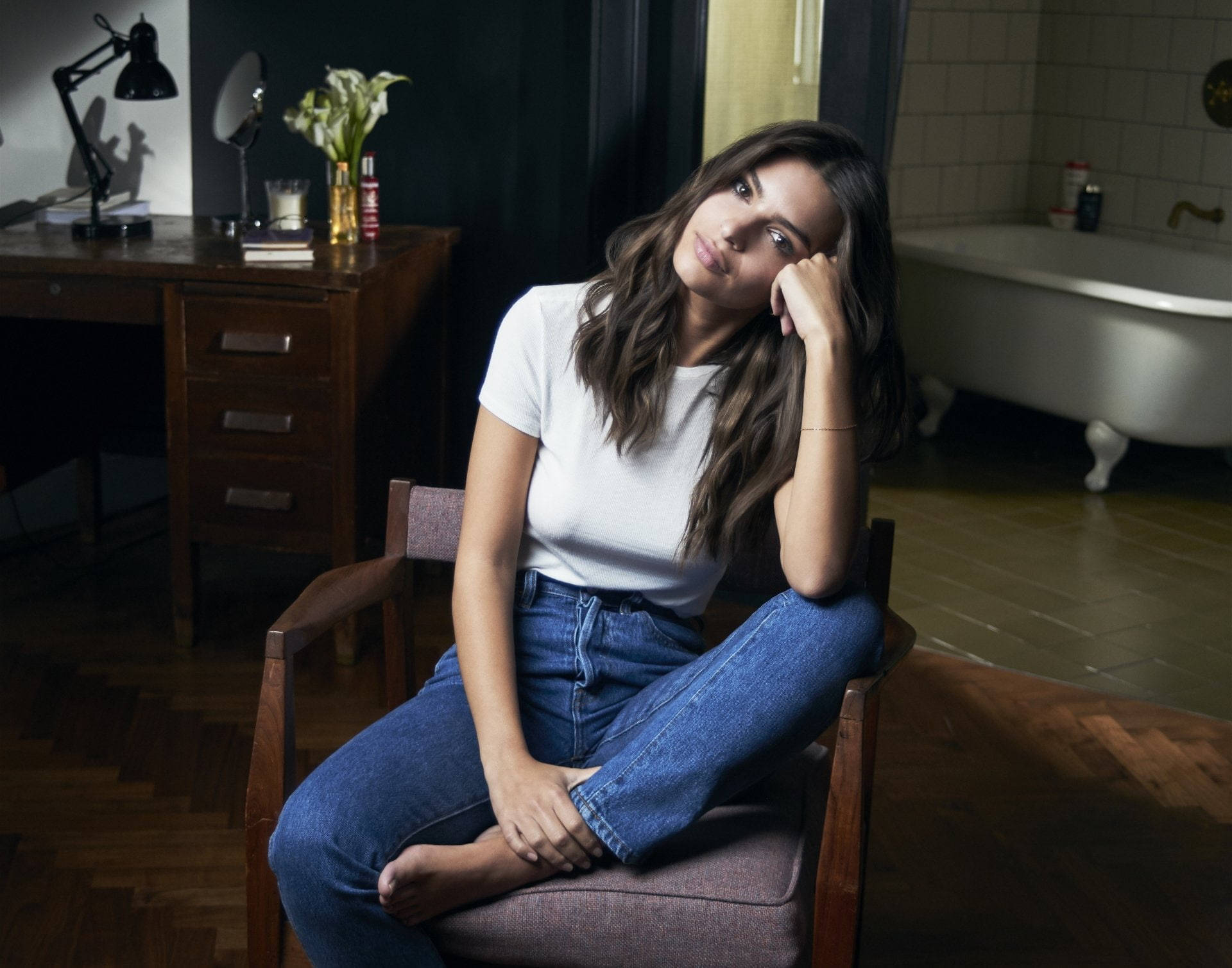 emily ratajkowski 桌布 壁紙 — 一位身穿白色T恤和牛仔褲的年輕女性，若有所思地坐在一個光線昏暗的復古浴室裡的木椅上，浴室裡有一個爪足浴缸和一張木桌。