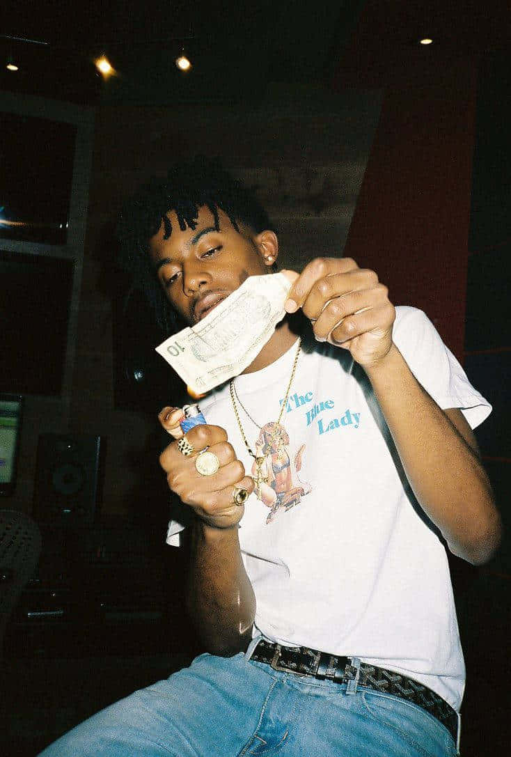 Playboi Carti HD 桌布 壁紙 — 一位身穿白色T恤和牛仔褲的年輕男子在一個光線昏暗、配有錄音室設備的房間裡手持一張燃燒的美元鈔票。