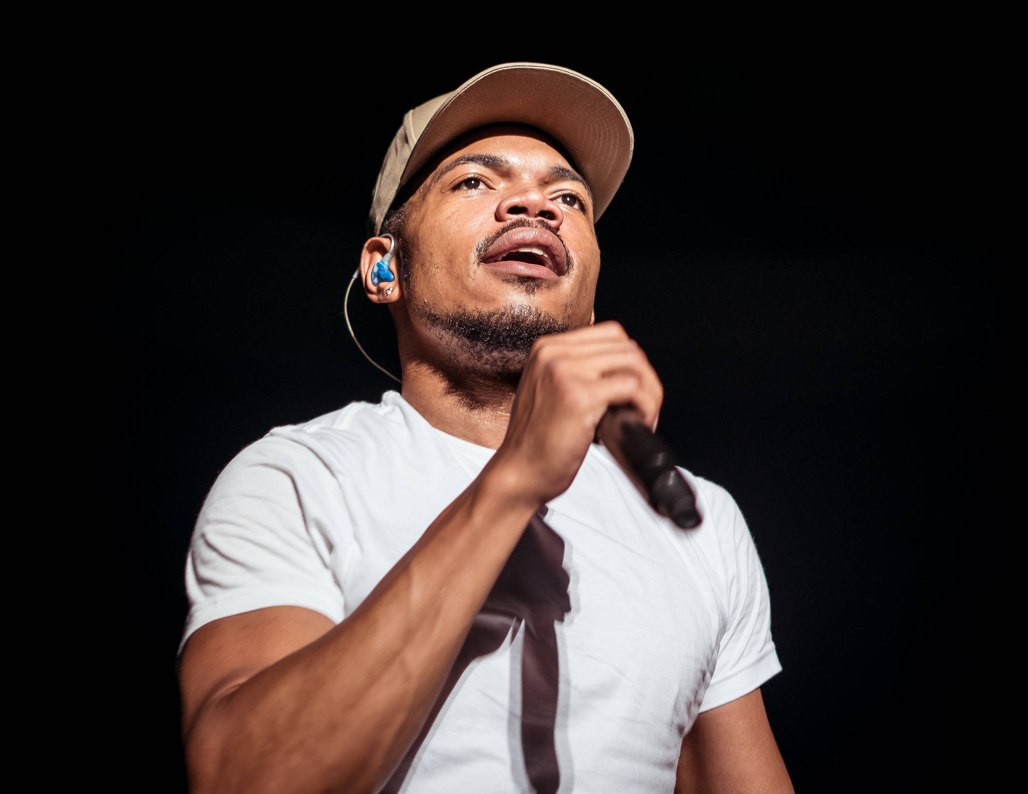 chance 桌布 壁紙 — 一位身穿白色T恤和米色帽子的男歌手，在黑色背景前對著麥克風充滿激情地演唱。