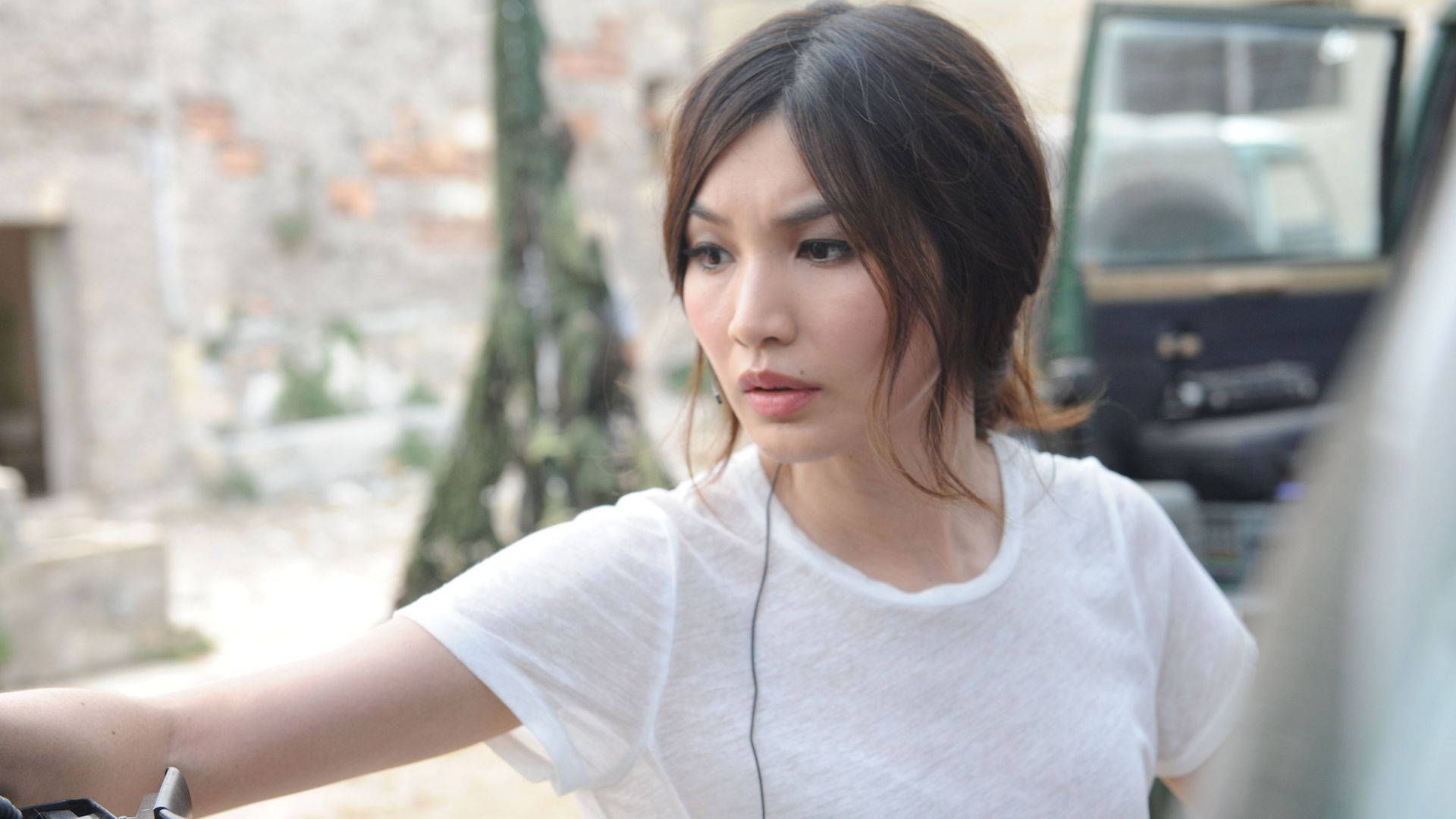 Gemma Chan 桌布 壁紙 — 一位身穿白色T恤的女人，戴著耳機，站在陽光普照的城市小巷裡一輛車附近，專注地望向前方。