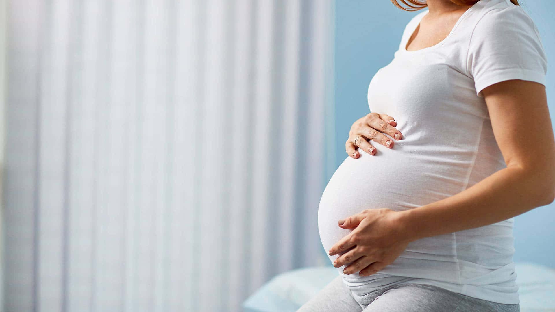 Pregnant 桌布 壁紙 — 一位身穿白色T恤的孕婦，在柔和的藍白背景下輕輕地抱著肚子。