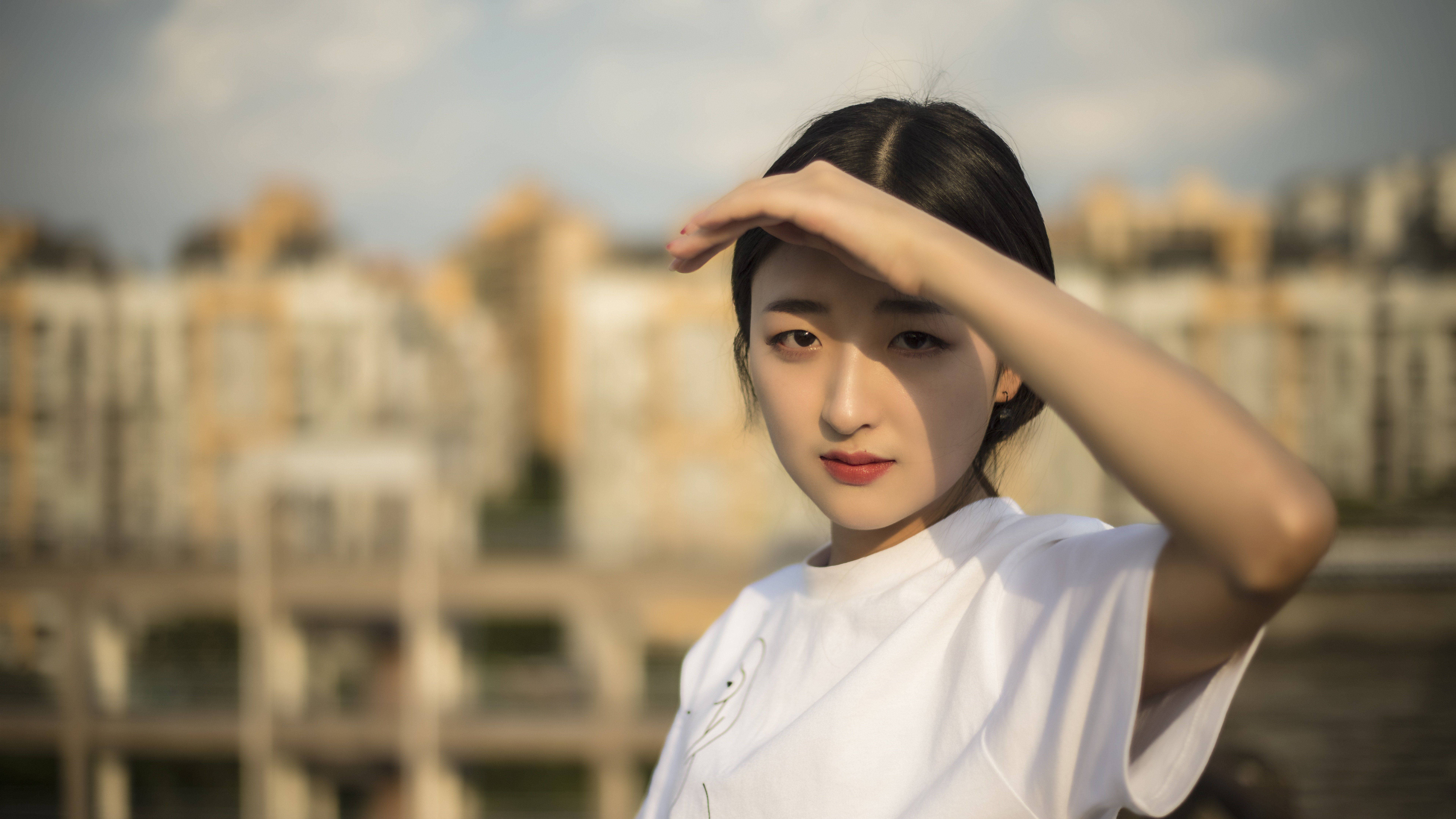 美女 桌布 壁紙 — 一位身穿白色T恤的年輕女性，在模糊的城市景觀背景下用手遮擋陽光。