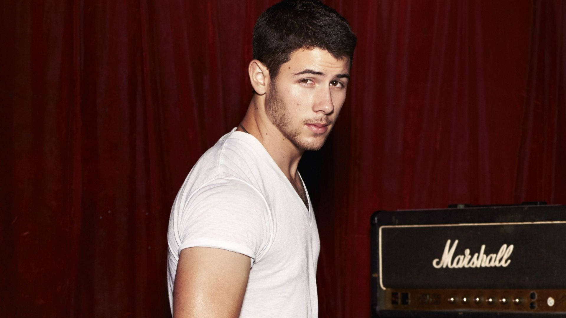 Nick Jonas 桌布 壁紙 — 一位身穿白色T恤的年輕男子站在深紅色窗簾旁的Marshall吉他擴音器旁邊。