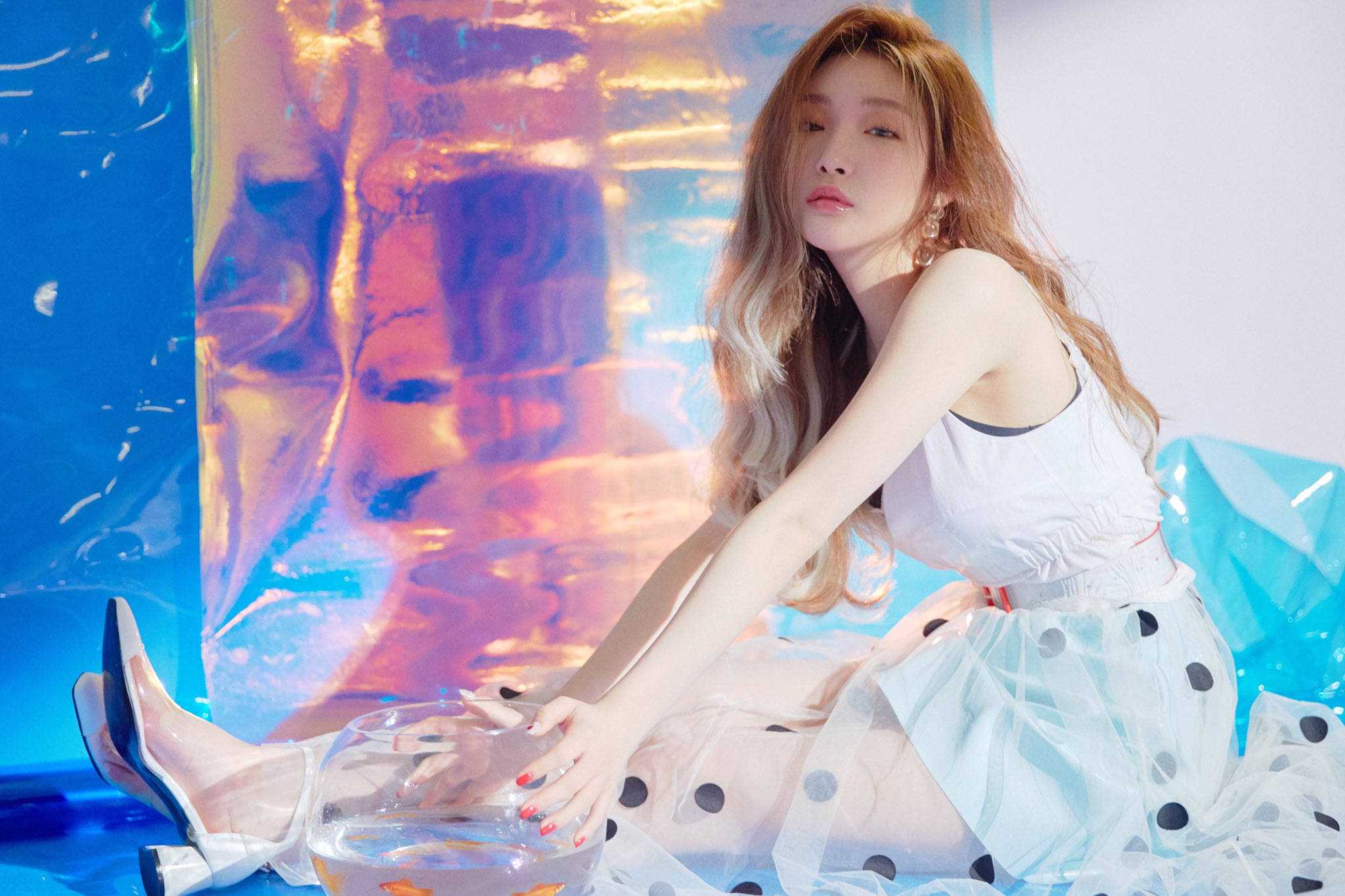 Chungha 桌布 壁紙 — 一位身穿白色上衣和圓點裙的女性，優雅地坐在魚缸旁，背景是充滿活力的藍色和粉紅色虹彩。