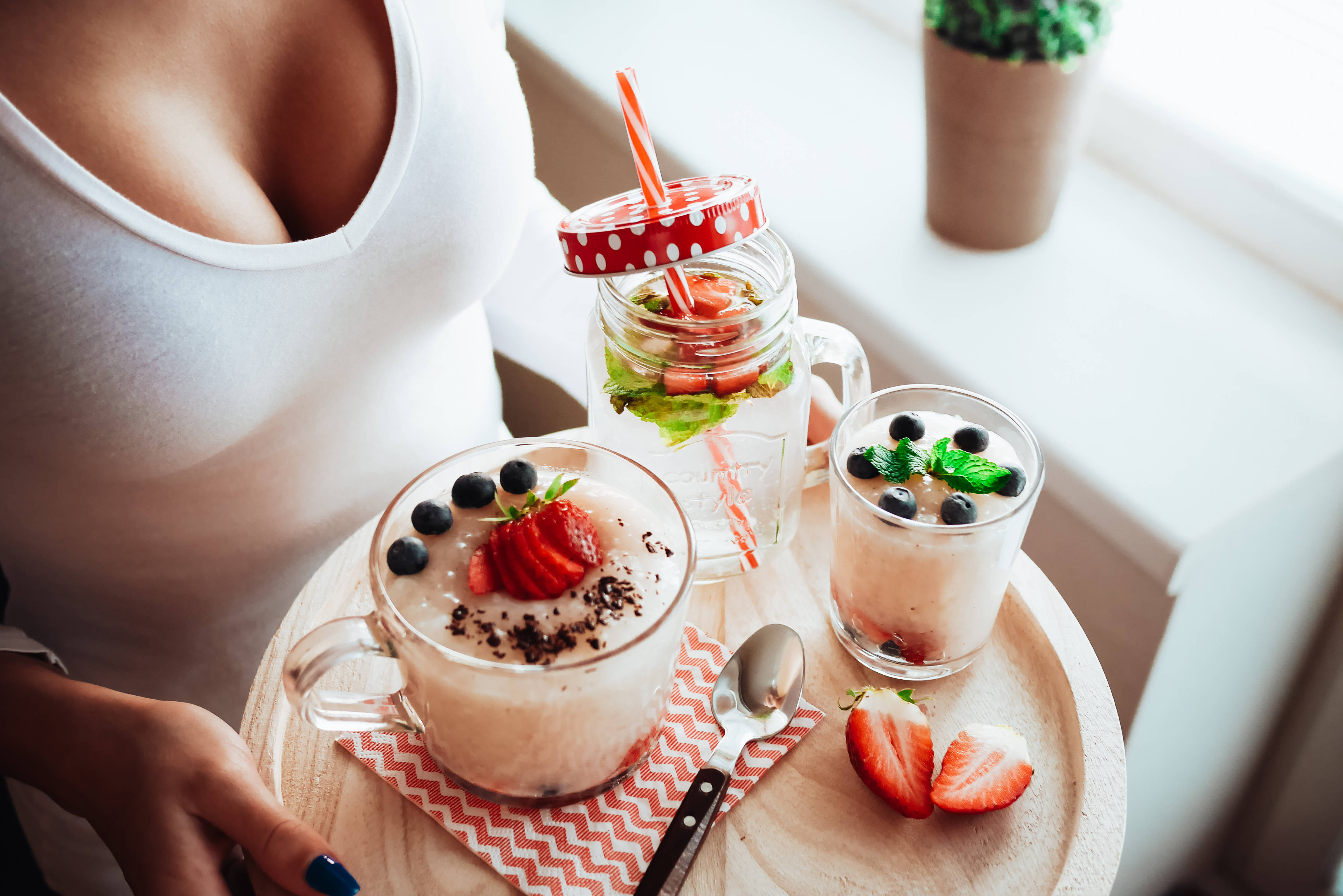 Fruits 桌布 壁紙 — 一位身穿白色上衣的女性在窗邊拿著一個木托盤，上面有三杯奶油般順滑的冰沙，頂部裝飾著草莓、藍莓和薄荷。