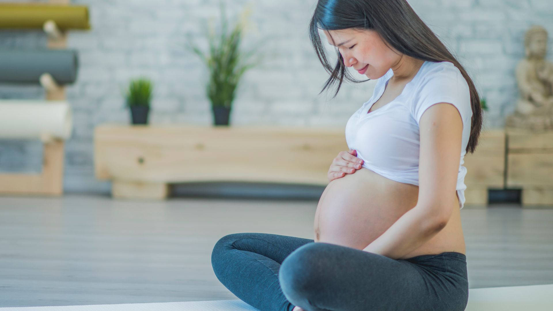 pregnancy 桌布 壁紙 — 一位身穿白色上衣的孕婦，在明亮的簡約工作室中盤腿坐在瑜伽墊上，輕輕地抱著肚子。