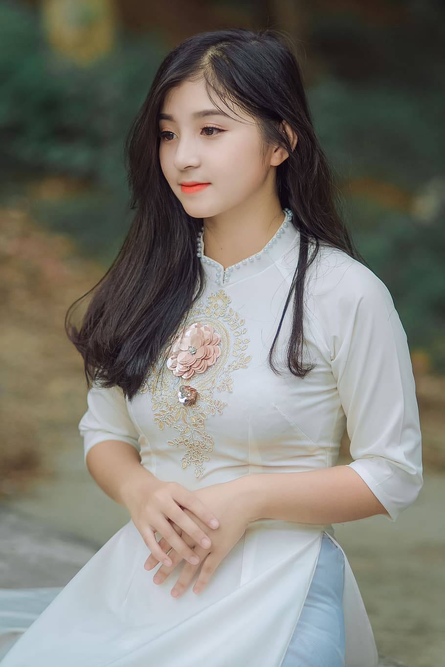 女孩 桌布 壁紙 — 一位身穿白色傳統刺繡花卉裙的年輕女性，優雅地坐在柔和模糊的自然環境中。
