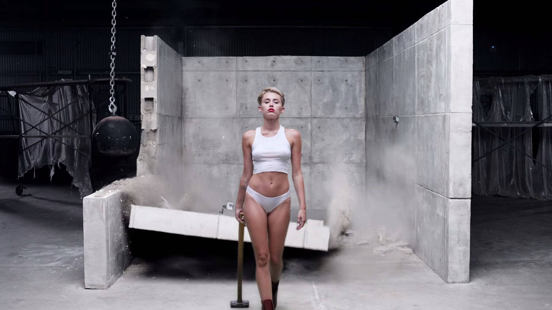 miley 桌布 壁紙 — 一位身穿白色內衣的女性自信地站在灰塵瀰漫的混凝土工業空間中，手持大錘。