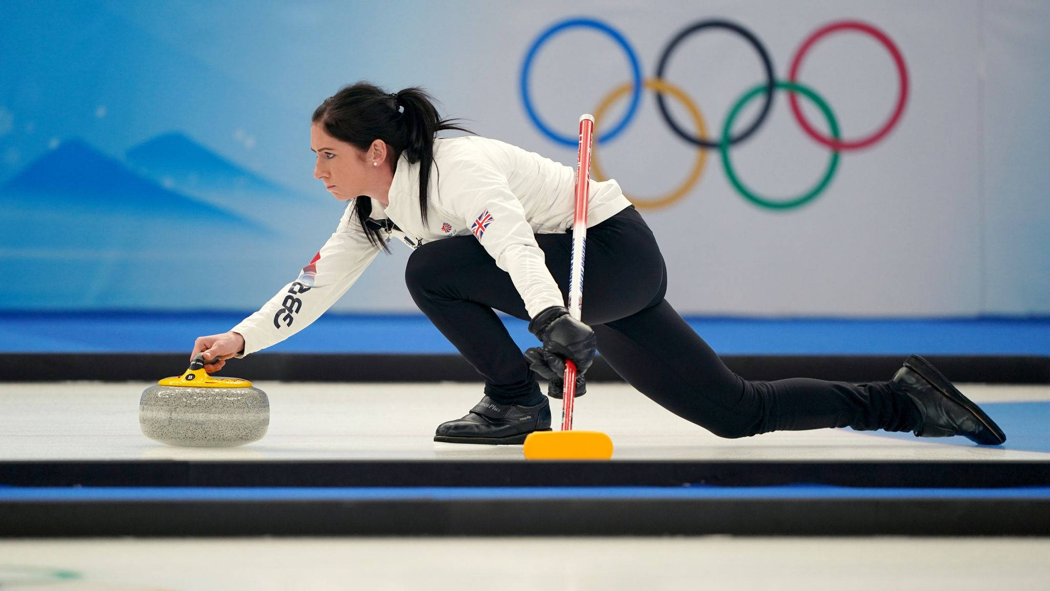 Olympics 桌布 壁紙 — 一位身穿白色制服、專注的女性冰壺運動員在冰面上推動花崗岩冰壺，背景可見奧運五環。
