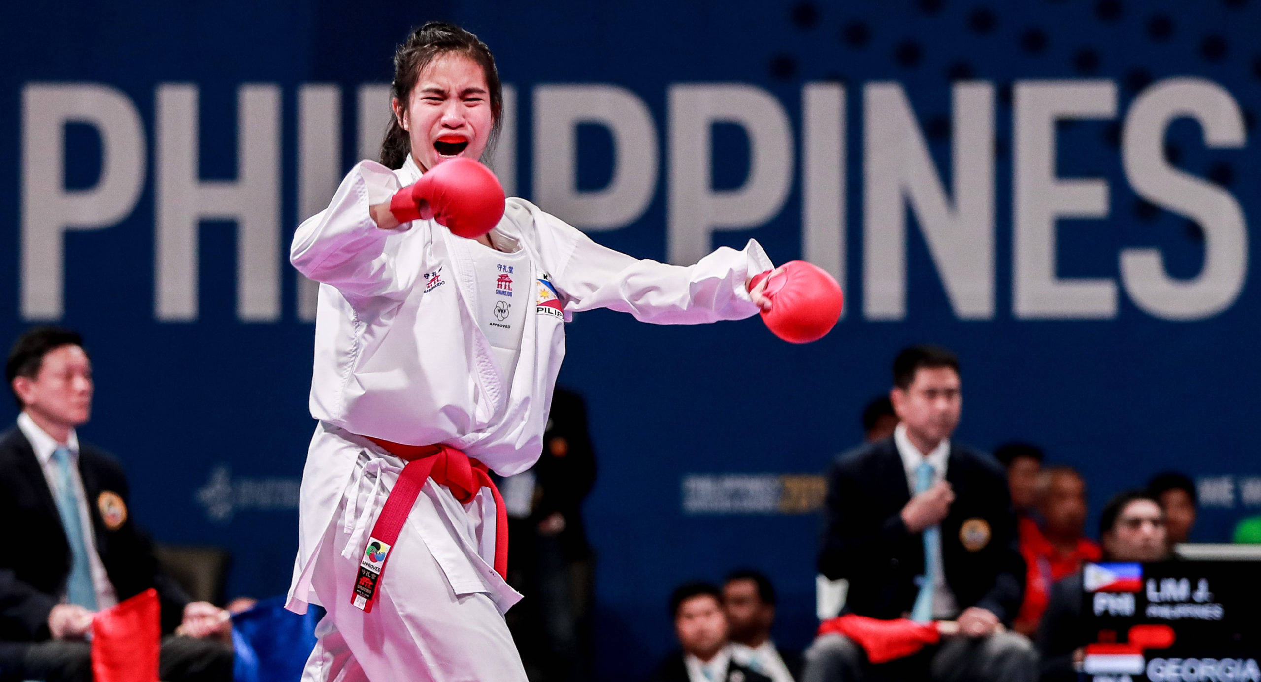Athletic 桌布 壁紙 — 一位身穿白色制服和紅色手套的女性跆拳道運動員在「PHILIPPINES」背景前進行激烈比賽時發出強力一拳。