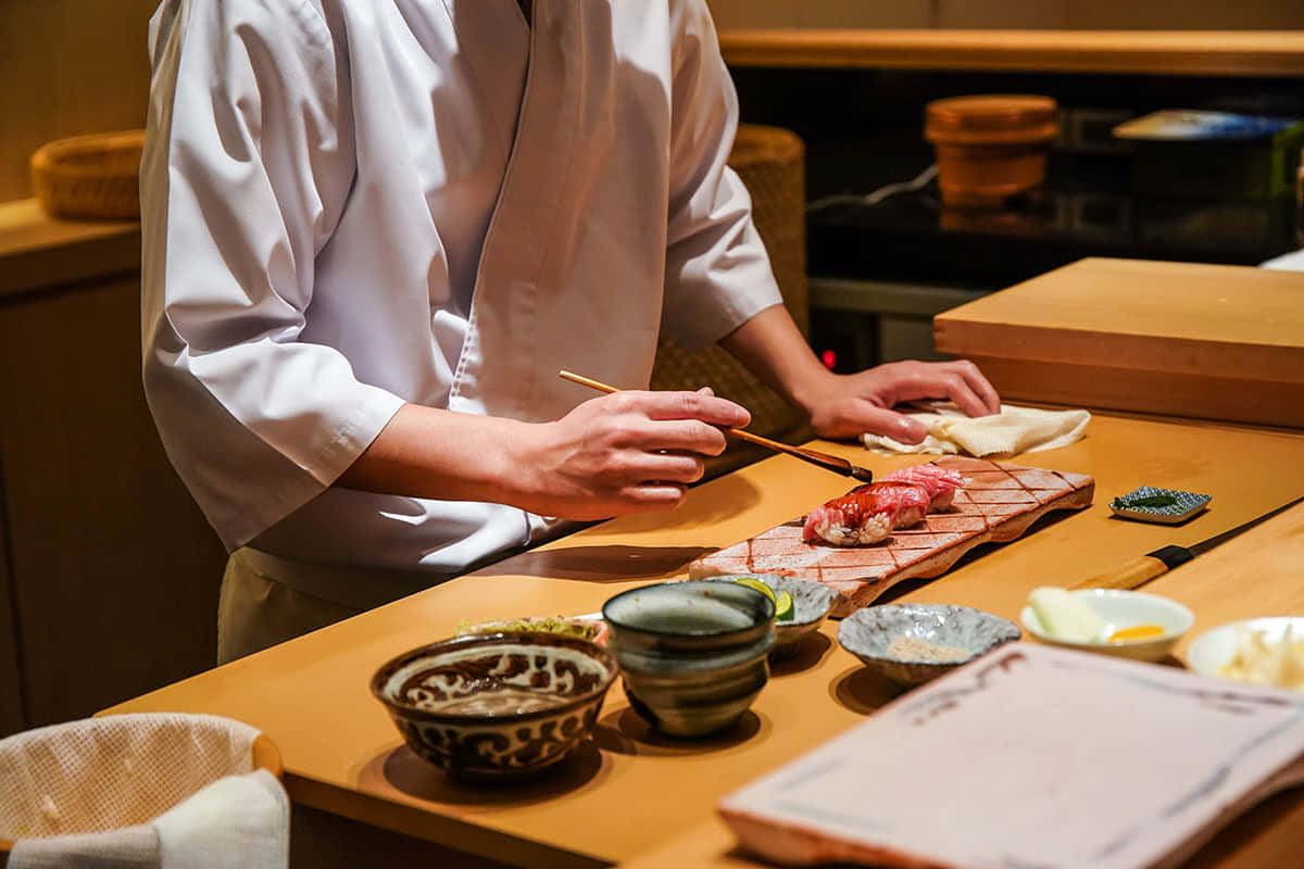 Tokyo Japan 桌布 壁紙 — 一位身穿白色制服的壽司師傅在溫暖燈光下的吧檯上，小心地將新鮮生魚片擺放在木板上。