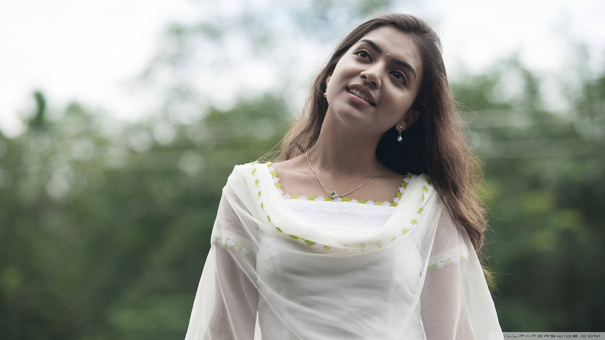 Indian 桌布 壁紙 — 一位身穿白色刺繡服裝的年輕女性，在模糊的綠色樹葉背景下，溫柔地微笑著向上凝視。