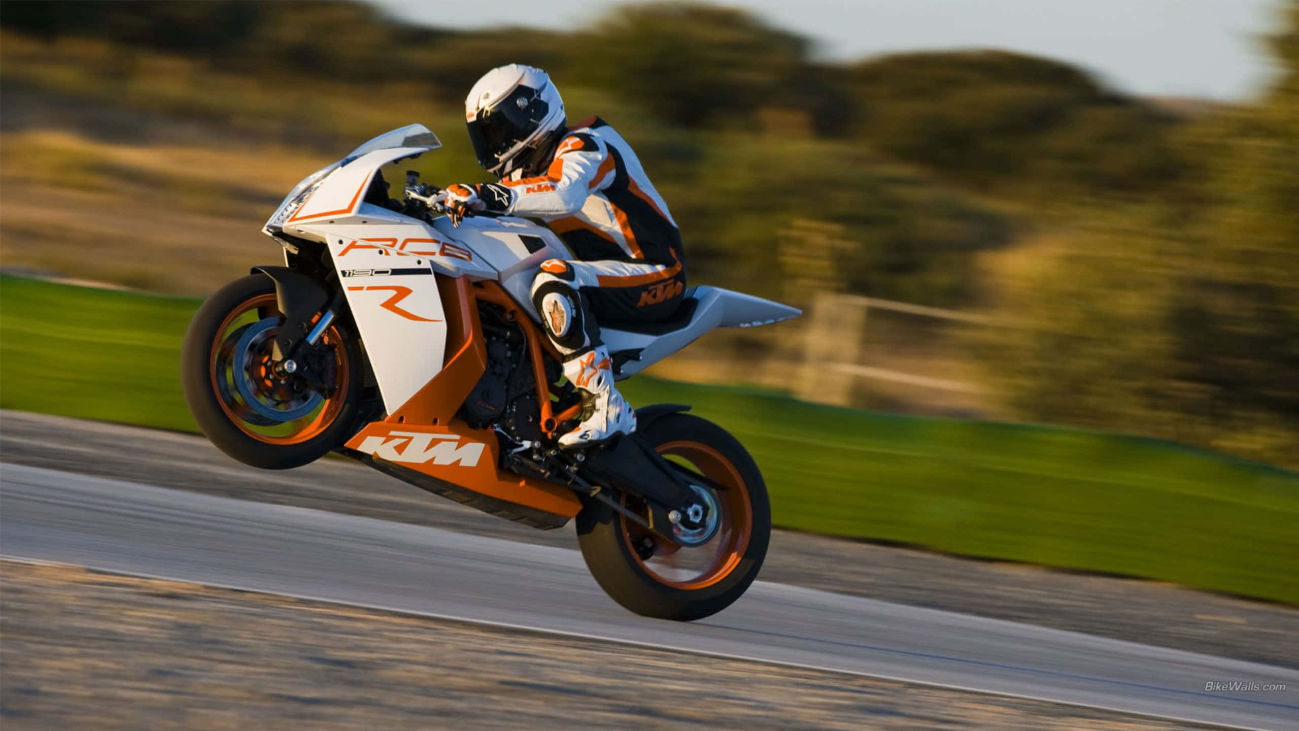 Ktm Bike 桌布 壁紙 — 一位身穿白色和橙色裝備的騎士，在陽光明媚的賽道上，騎著白色和橙色的KTM RC8R摩托車傾斜入彎，背景是模糊的綠色植物。