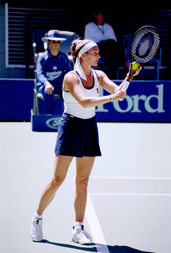 Tennis 桌布 壁紙 — 一位身穿白色和海軍藍裝備的女網球選手在陽光明媚的硬地球場上準備正手擊球。