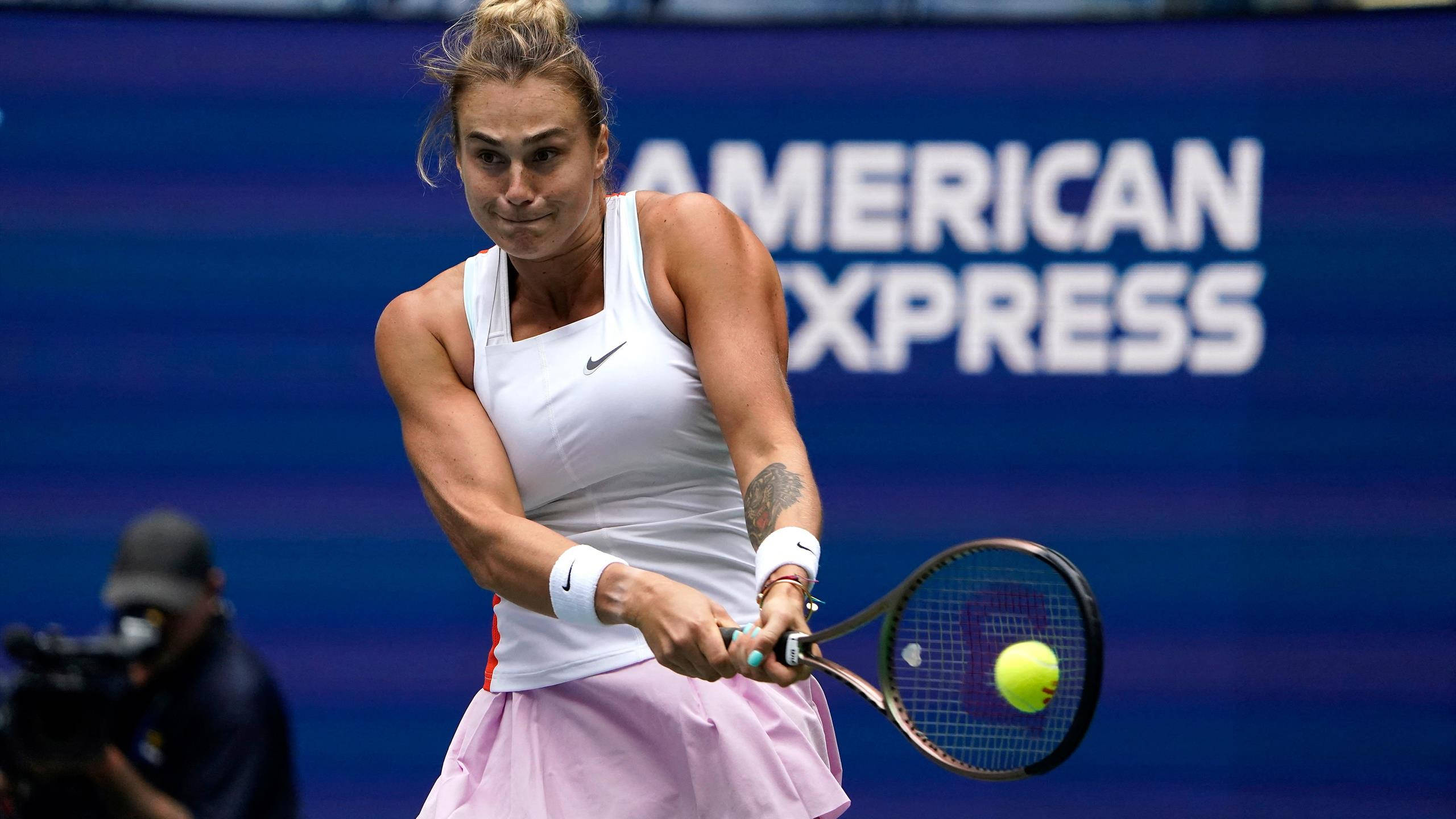 Aryna Sabalenka 桌布 壁紙 — 一位身穿白色和粉紅色裝備的女網球選手，在藍色美國運通背景前揮拍擊打黃色球。