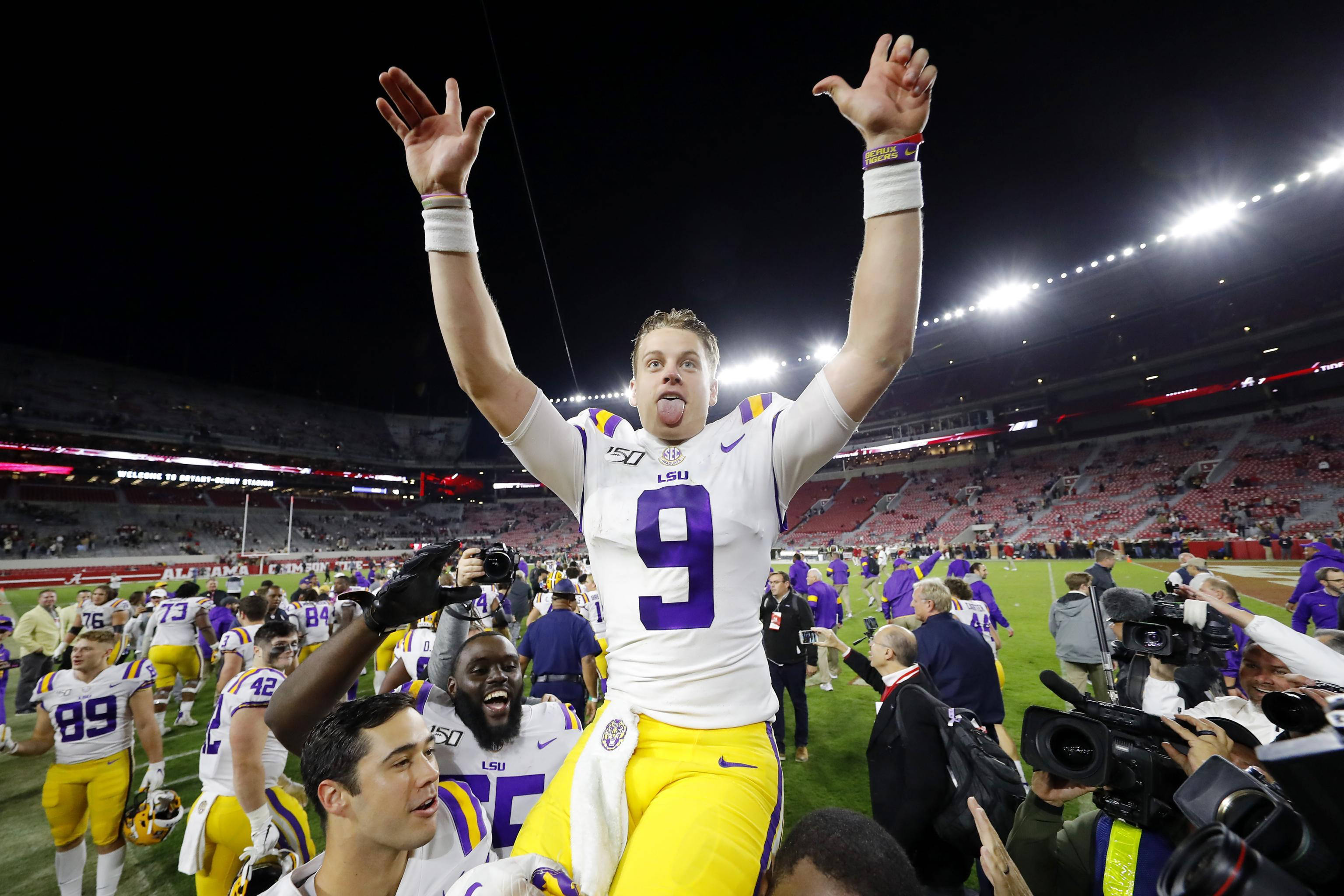 Joe Burrow 桌布 壁紙 — 一位身穿白色和紫色9號球衣的LSU美式足球員，在夜間燈光球場上高舉雙臂、伸出舌頭歡呼慶祝。