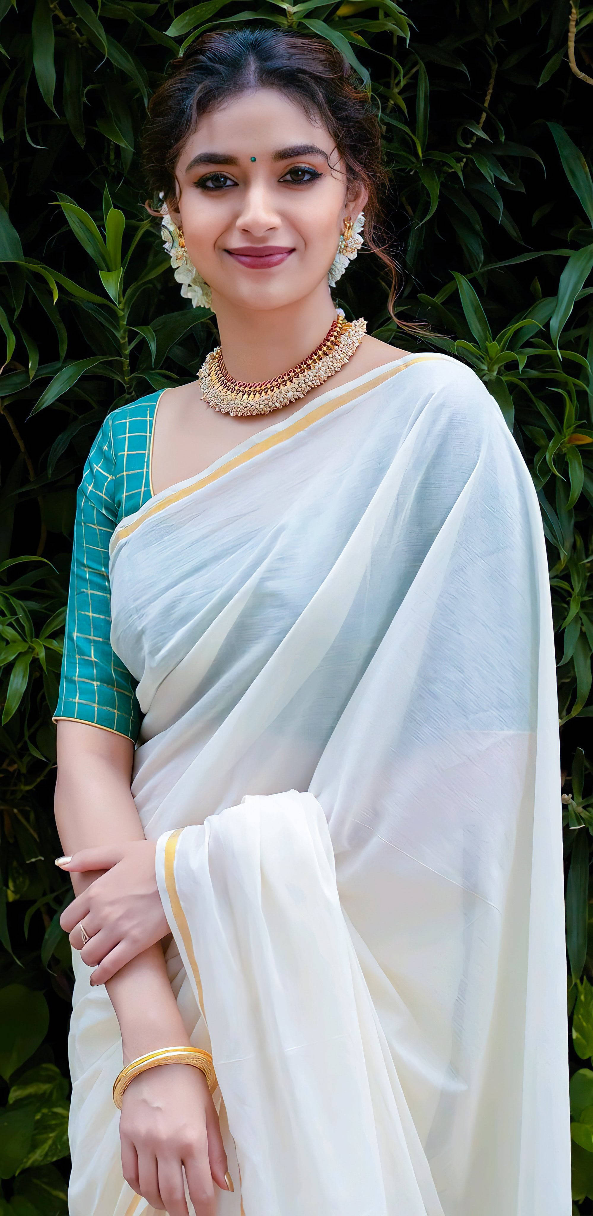 telugu 桌布 壁紙 — 一位身穿白色和藍綠色紗麗、帶有金色點綴的女性，在茂密的綠色樹葉背景下優雅地微笑。