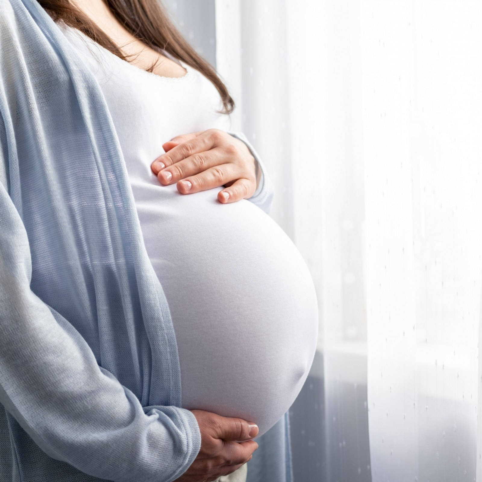 Pregnant 桌布 壁紙 — 一位身穿白色和藍色的孕婦在明亮的窗邊輕柔地抱著肚子，窗簾是薄紗的。