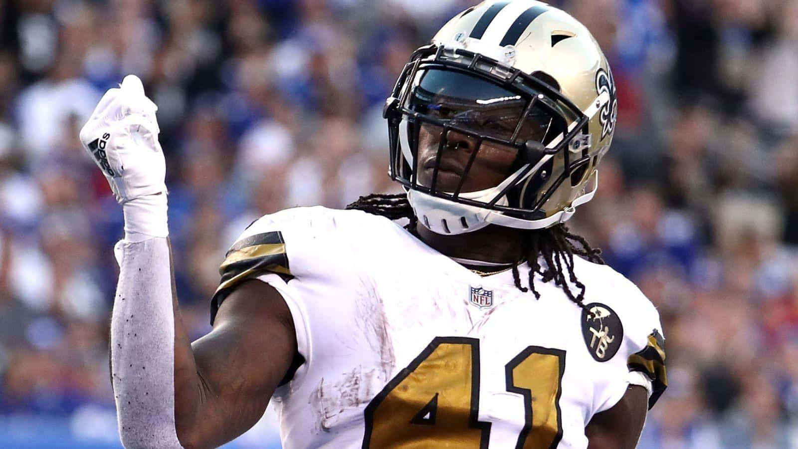 Alvin Kamara 桌布 壁紙 — 一位身穿白色和金色球衣的紐奧良聖徒隊美式足球員在陽光明媚的球場上舉起拳頭慶祝勝利，背景是模糊的人群。