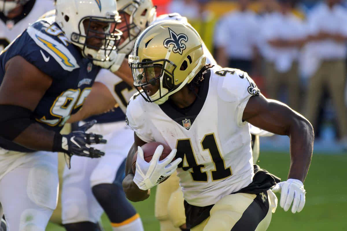 Alvin Kamara 桌布 壁紙 — 一位身穿白色和金色球衣的紐奧良聖徒隊美式足球員在陽光照耀的球場上帶球衝刺，身後有閃電隊防守球員追趕。