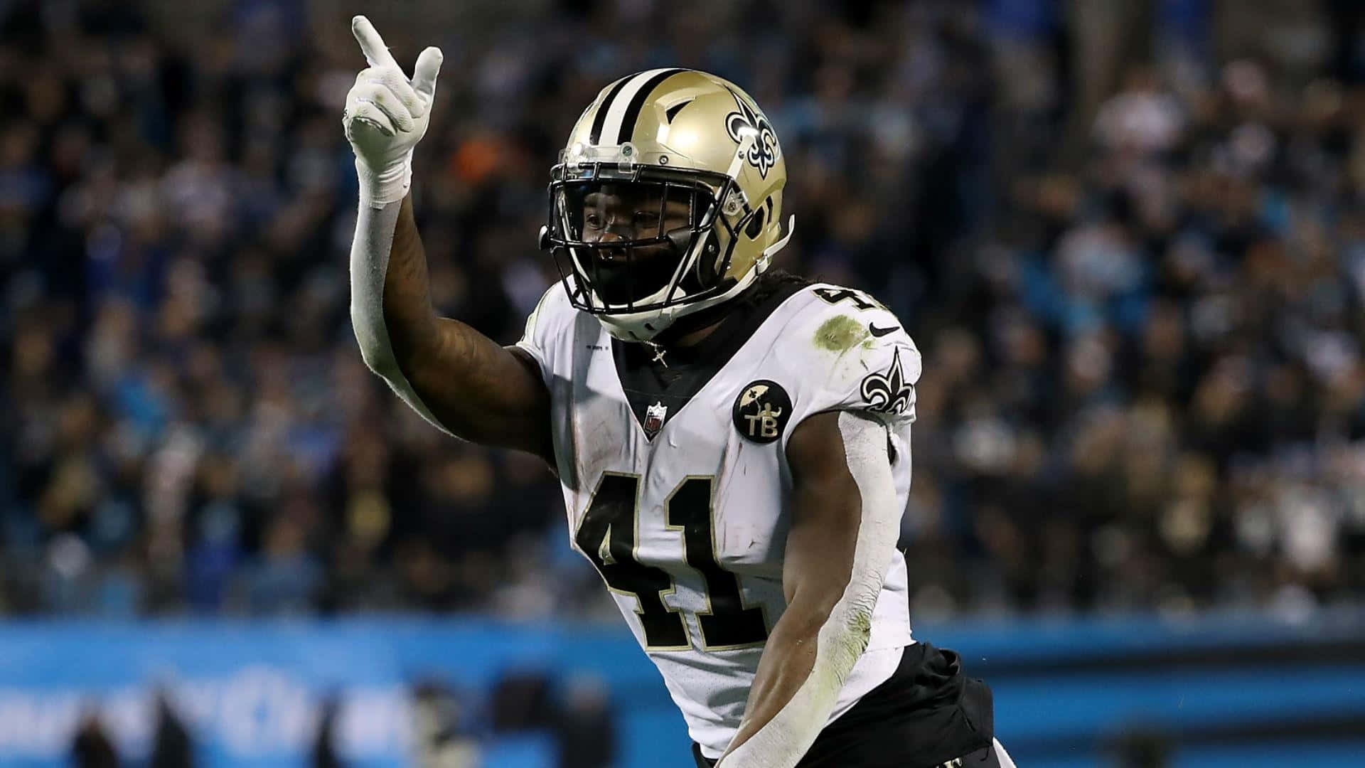 Alvin Kamara 桌布 壁紙 — 一位身穿白色和金色球衣的紐奧良聖徒隊美式足球員在夜間比賽中於球場燈光下舉手慶祝。