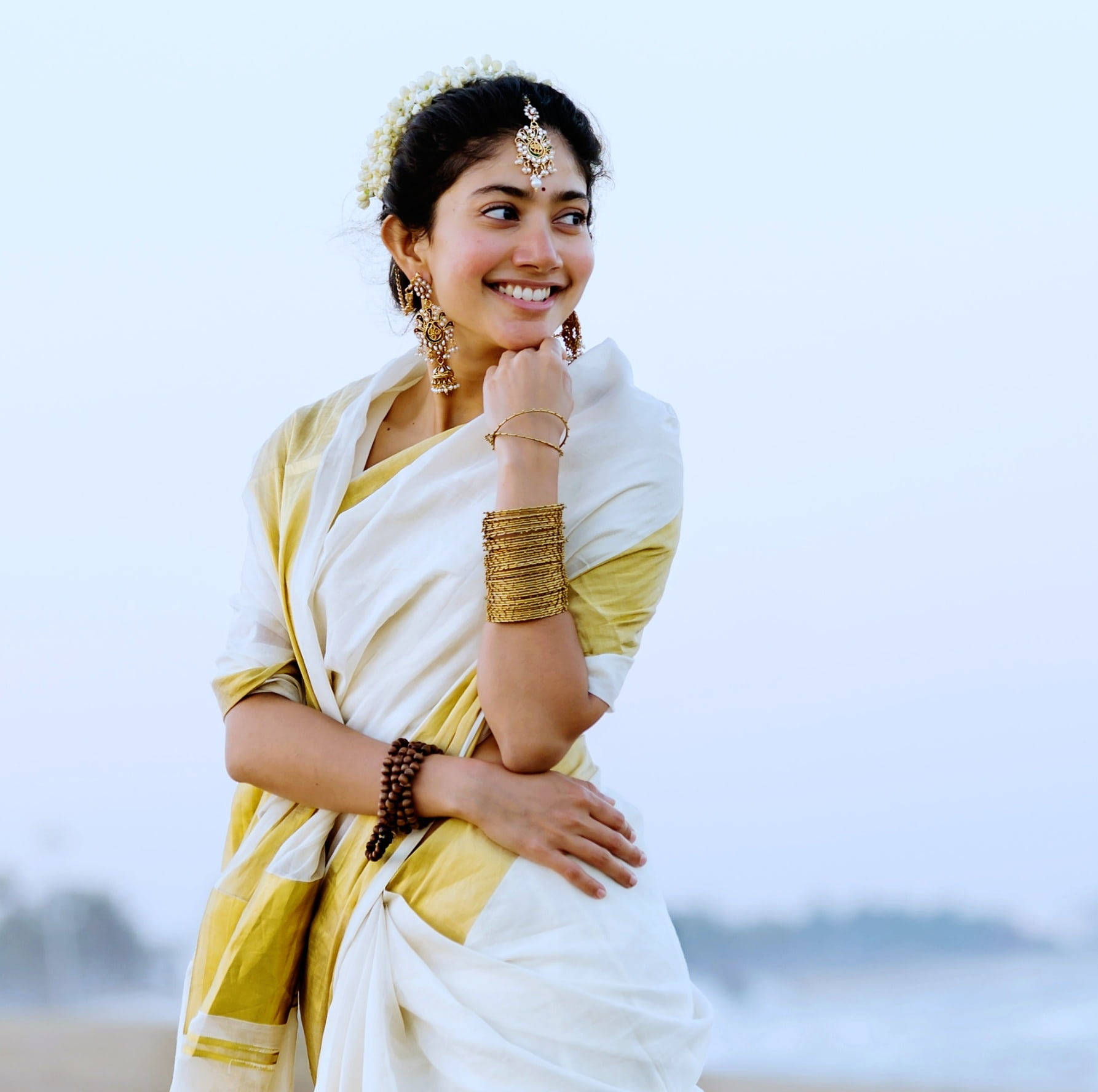 Sai Pallavi 桌布 壁紙 — 一位身穿白色和金色紗麗的光彩照人的女性在霧濛濛的海灘上微笑，背景是海浪。