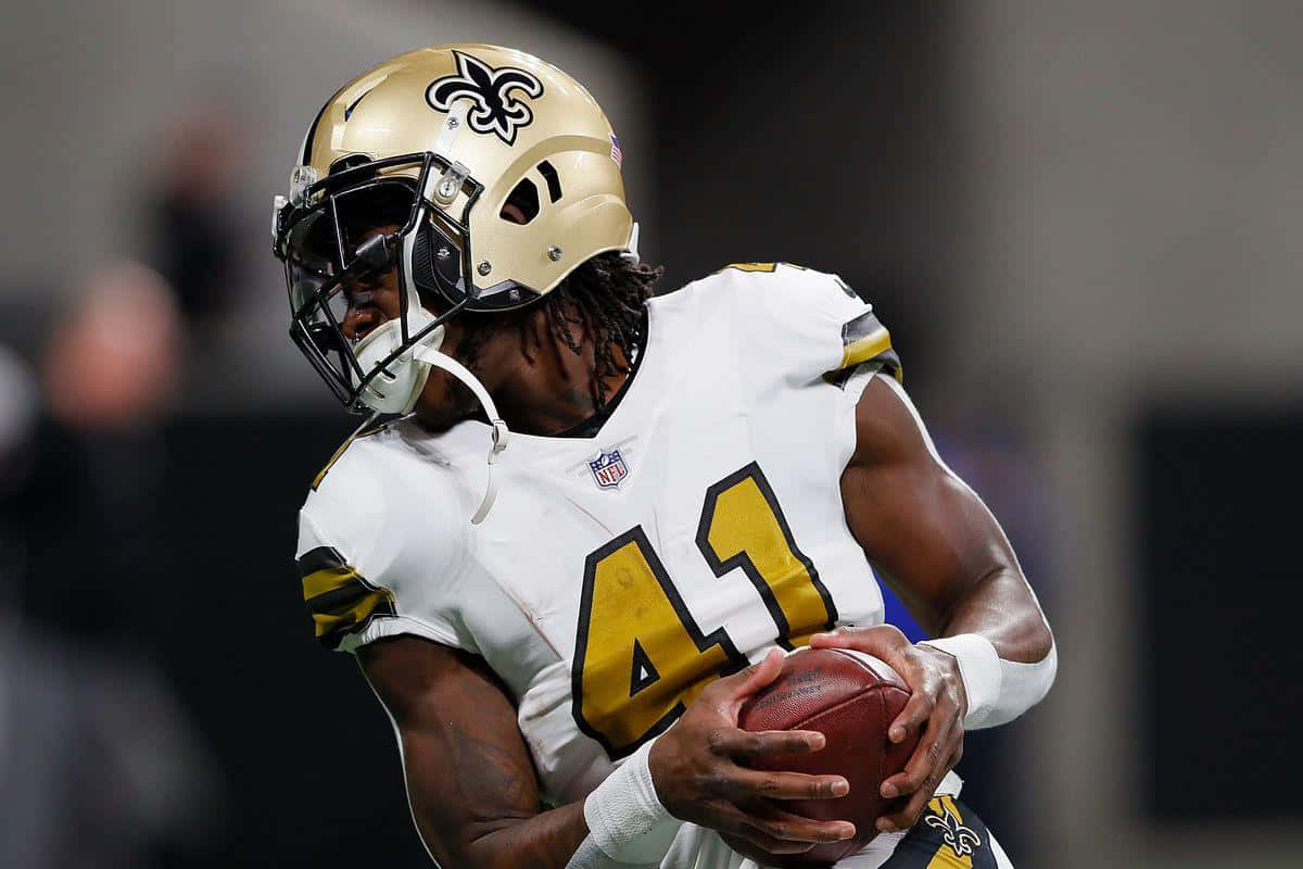 Alvin Kamara 桌布 壁紙 — 一位身穿白色和金色裝備的紐奧良聖徒隊美式足球員在比賽中專注地緊握著球。
