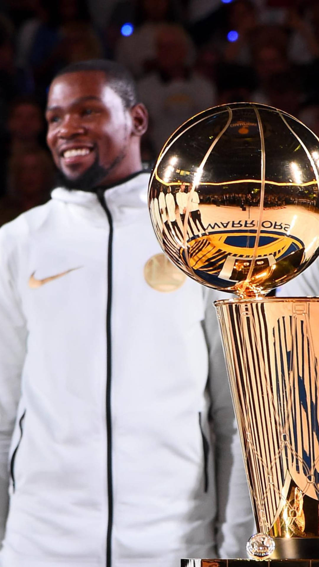 Trophy 桌布 壁紙 — 一位身穿白色外套、面帶微笑的籃球運動員站在閃閃發光的金色NBA冠軍獎盃後面，獎盃反射著場館的燈光。
