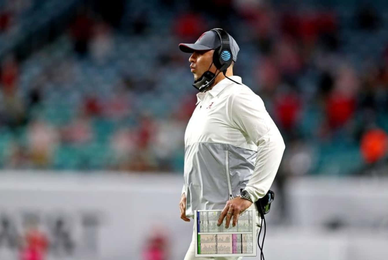 Coach 桌布 壁紙 — 一位身穿白色外套和耳機的足球教練在比賽期間專注地站在邊線，身後是模糊的體育場座位。