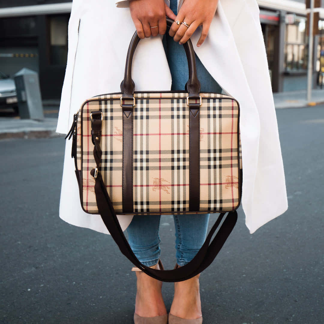 Burberry 桌布 壁紙 — 一位身穿白色外套的人，在城市街道上拿著一個經典的米色格紋 Burberry 公事包，配有棕色皮革帶。