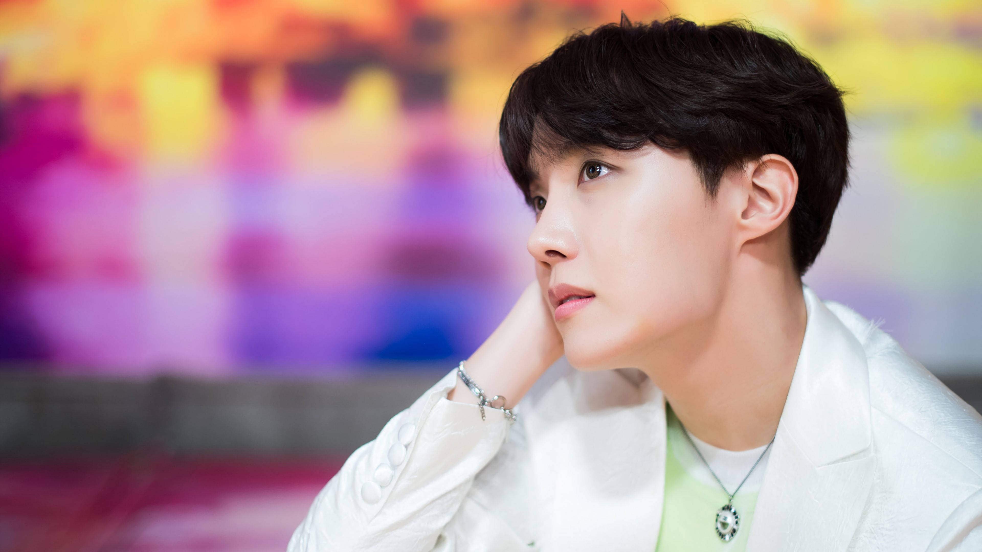 BTS 桌布 壁紙 — 一位身穿白色外套的年輕男子，在充滿活力的模糊彩虹背景下若有所思地凝視。