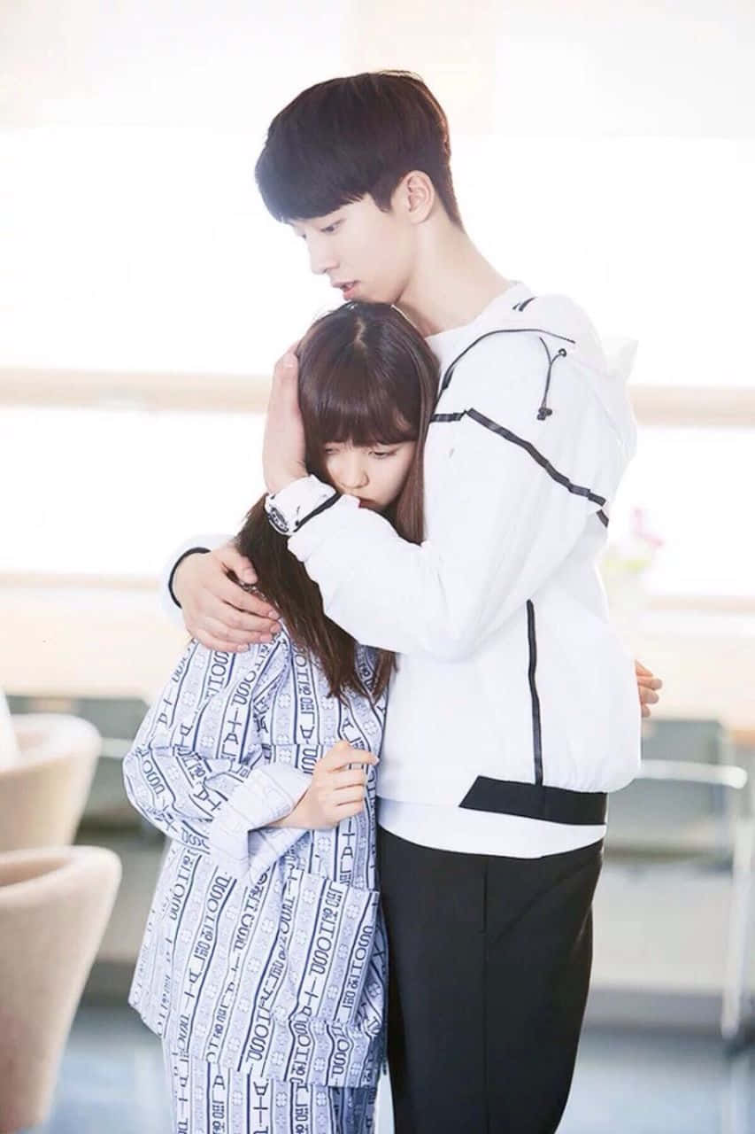 Couples 桌布 壁紙 — 一位身穿白色外套的年輕男子輕輕擁抱一位穿著圖案睡袍的女孩，她將頭靠在他的胸膛上，身處柔和光線的室內空間。