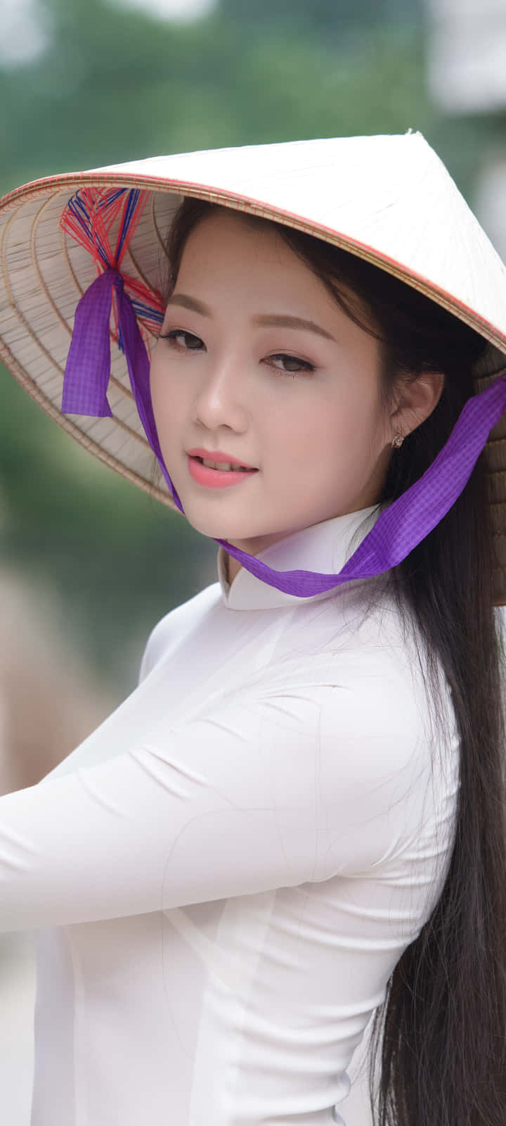 Vietnam 桌布 壁紙 — 一位身穿白色奧黛和傳統錐形帽（帶紫色絲帶）的年輕女性，在模糊的綠色背景下溫柔地凝視。