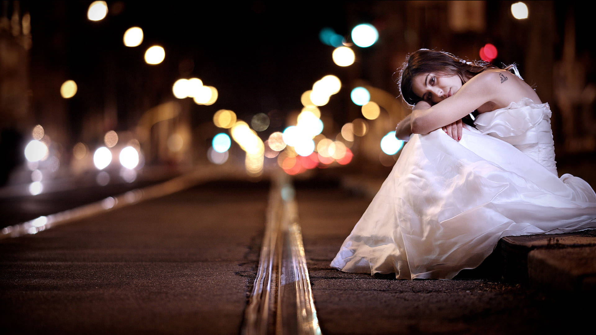 婚紗 桌布 壁紙 — 一位身穿白色婚紗的女性夜晚獨自坐在城市街道上，被交通中閃爍的散景燈光包圍。