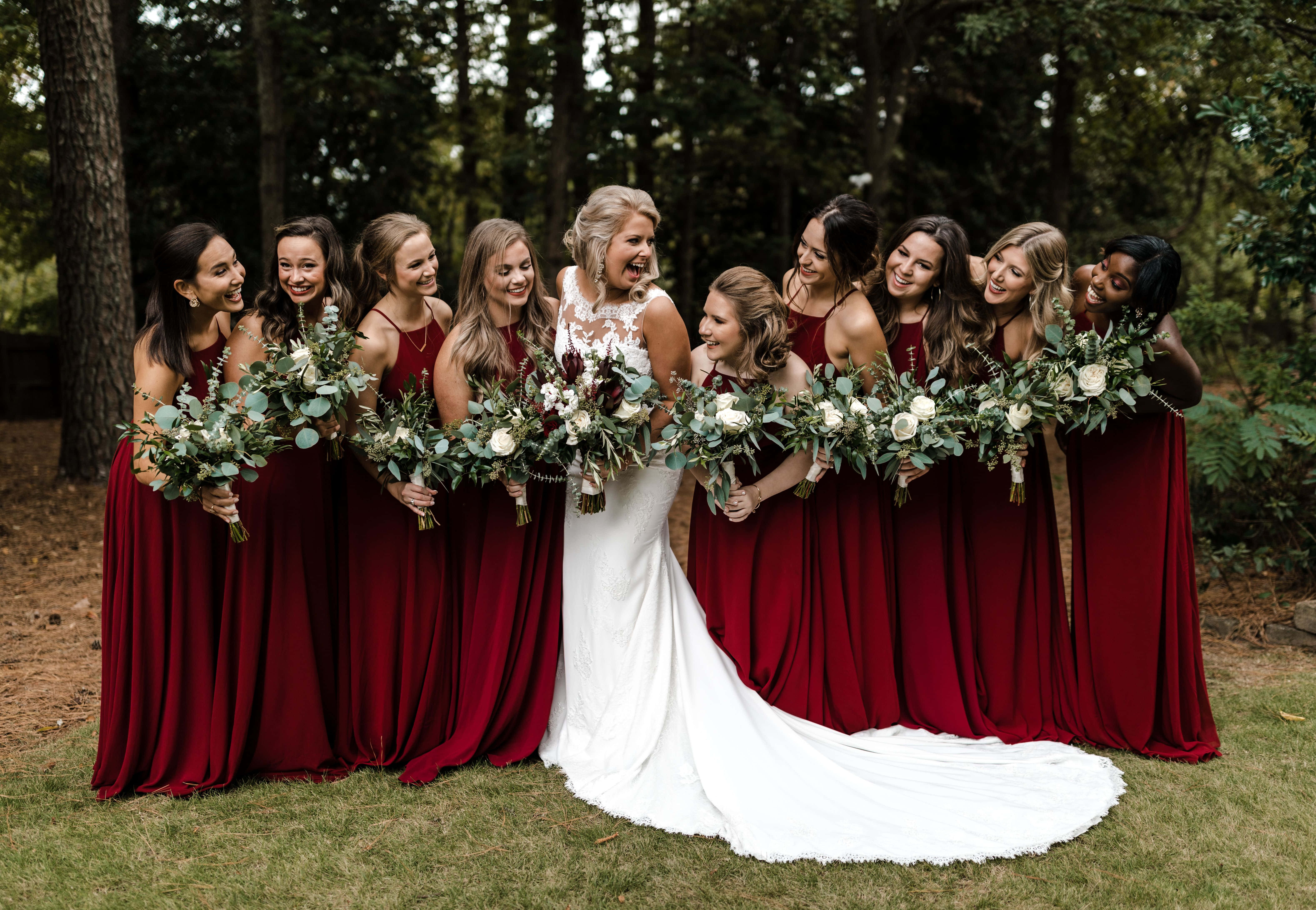 Bridesmaids 桌布 壁紙 — 一位身穿白色婚紗的快樂新娘，與身穿酒紅色禮服的伴娘們在森林裡手持花束歡笑。